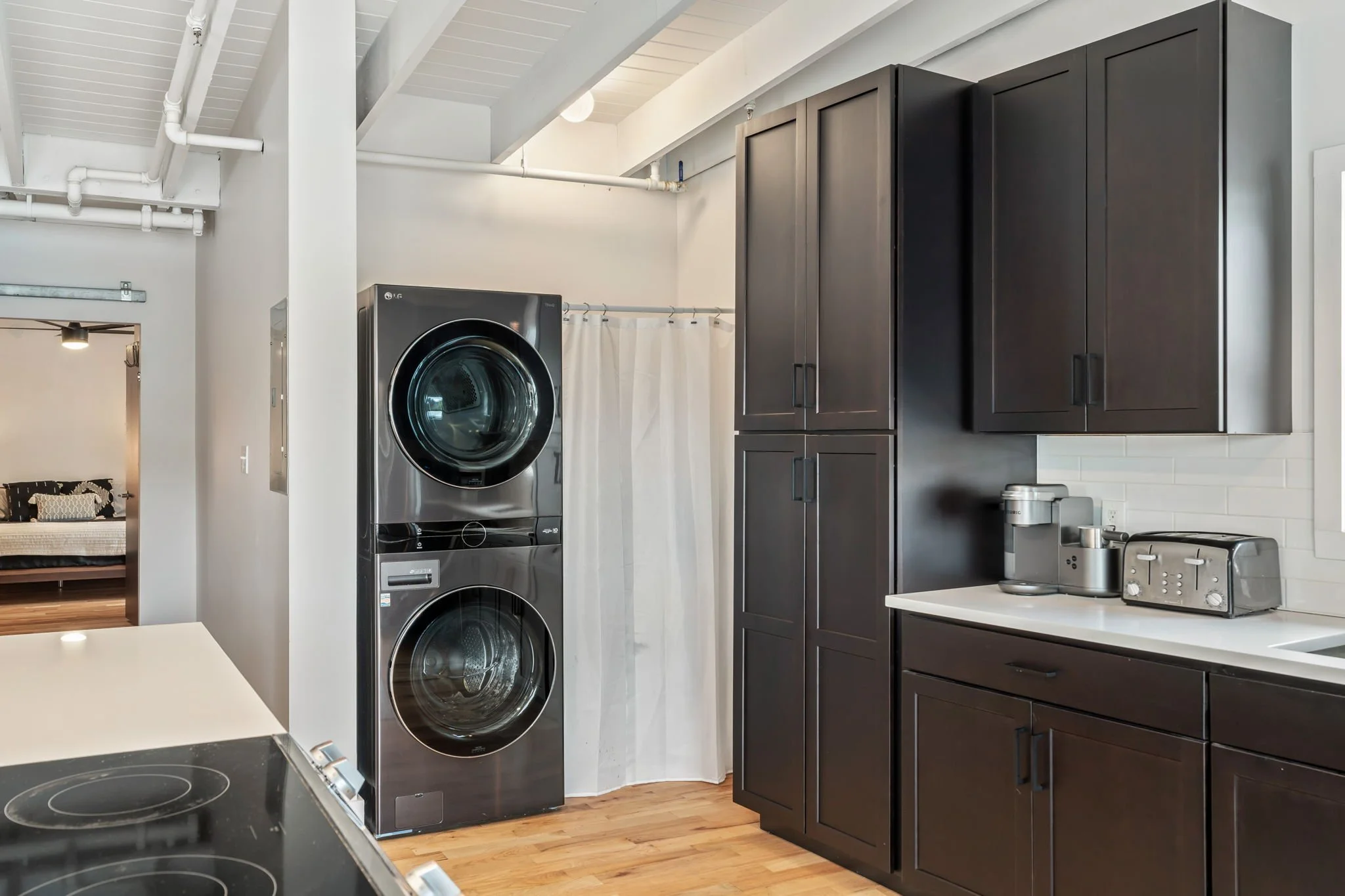 Laundry area with stacked black washer and dryer, dark brown kitchen cabinets, white countertop with a toaster and coffee machine, wooden floor, white wall, and curtain covering a doorway.