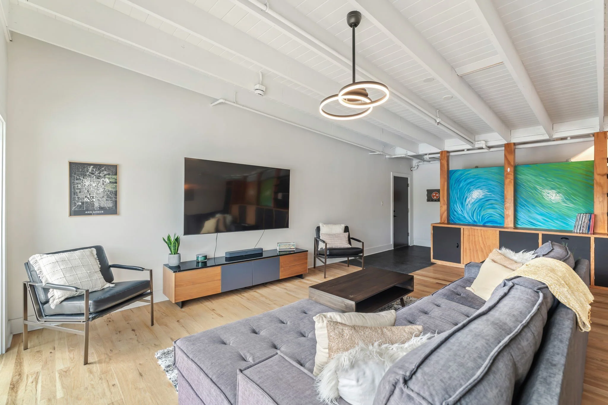 Modern living room with gray sectional sofa, black armchair, wooden coffee table, wall-mounted TV, abstract wall art, and artwork resembling ocean waves, with white walls, wooden flooring, and a unique circular pendant light.