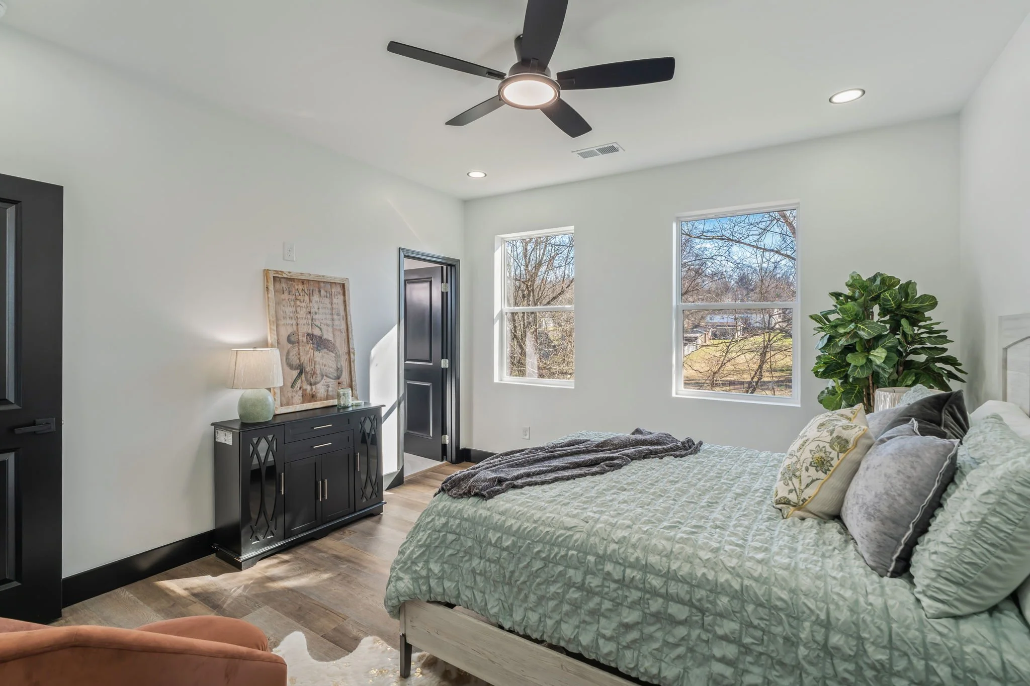 A bright bedroom with white walls, two windows showing trees outside, a black ceiling fan, a white bed with green and gray pillows, a black dresser with a lamp and decorative picture, a plant in the corner, and animal print on the wall.