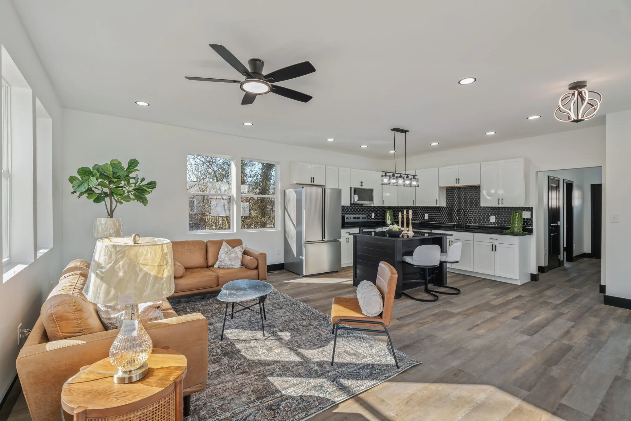 Open-concept living room and kitchen with white walls, hardwood floors, brown leather sofa, beige chair, coffee table, black kitchen island, white cabinetry, stainless steel refrigerator, and decorative lighting fixtures.