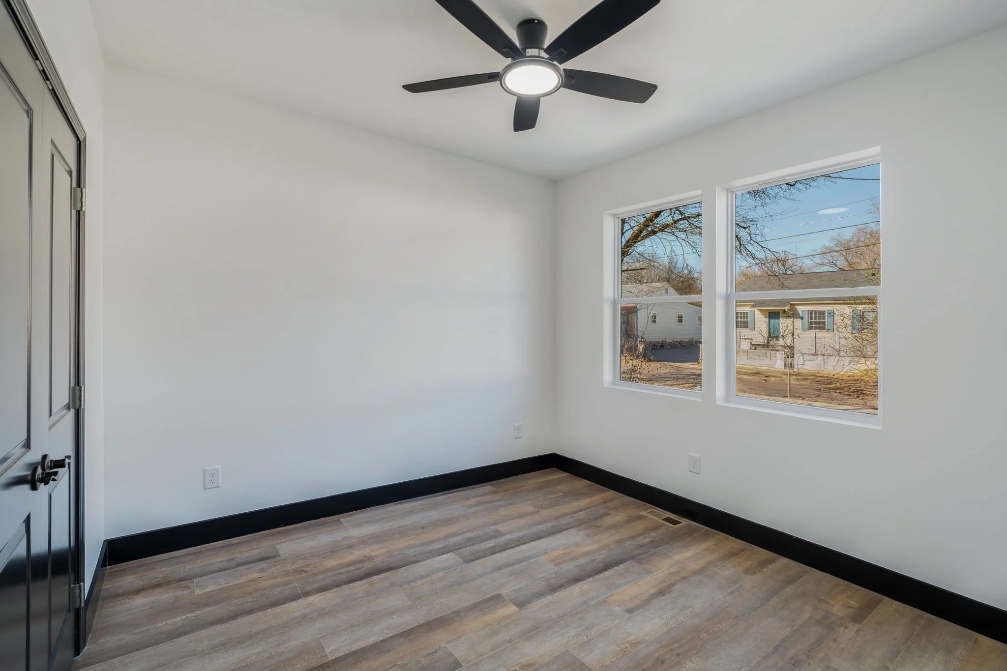 Empty room with white walls, hardwood flooring, two windows showing houses outside, a ceiling fan, and black trim along the floor.