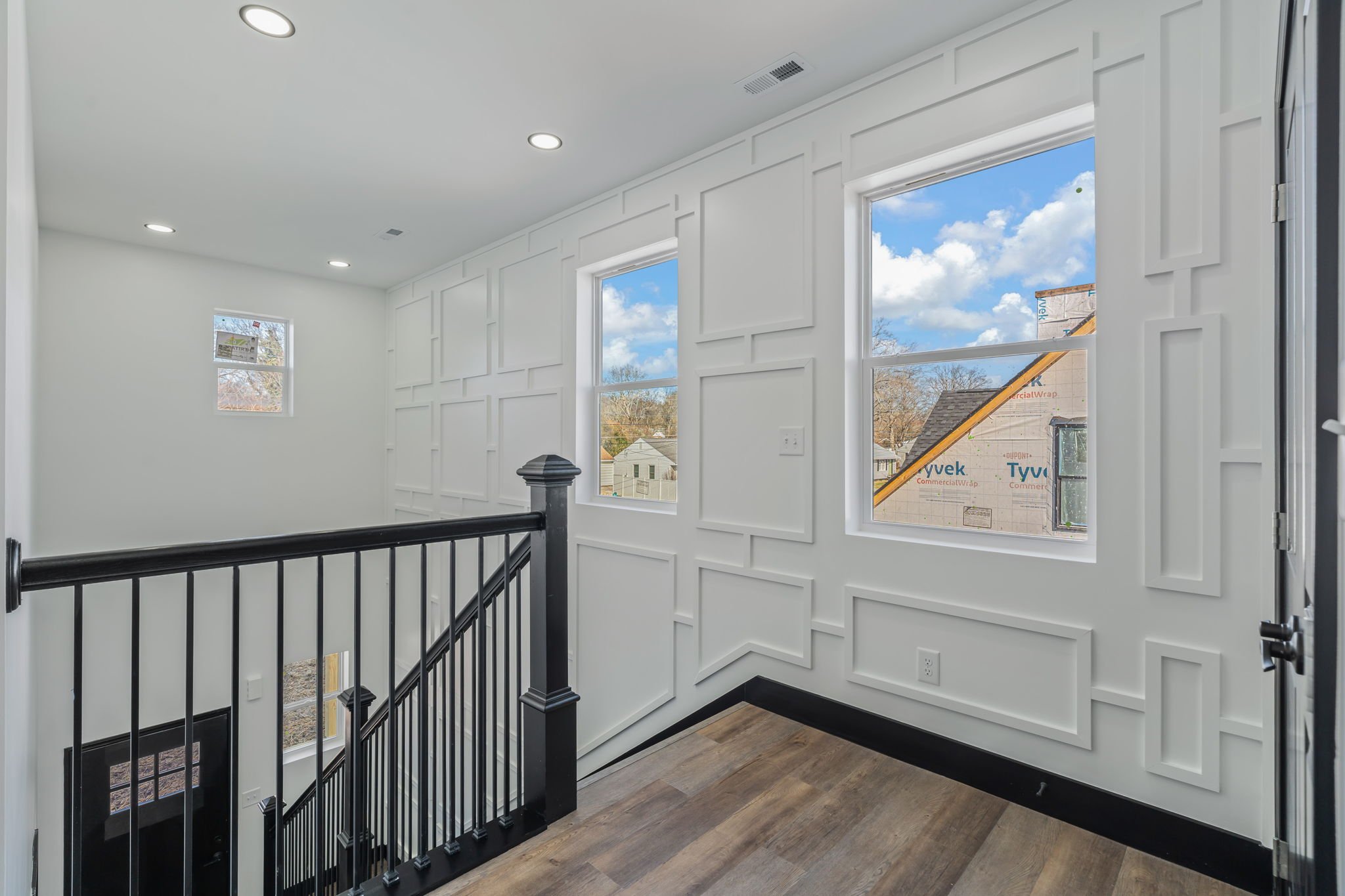 Interior view of a house under construction with white paneled walls, three windows showing blue sky and neighborhood, black railing, and wooden floor.