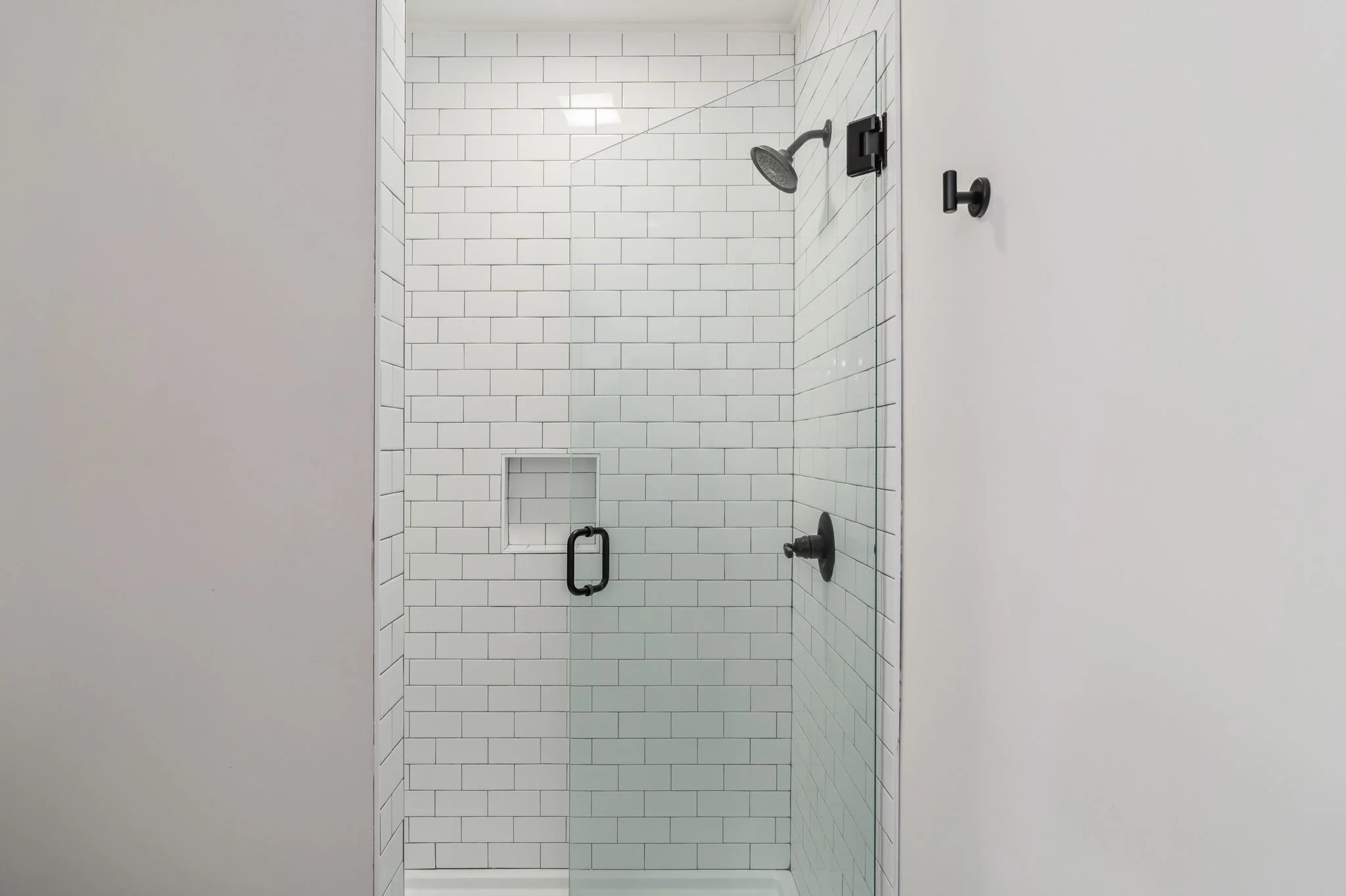 Modern glass-enclosed shower with white subway tile walls, a black showerhead, a black handle, a black control knob, and a built-in niche for toiletries.