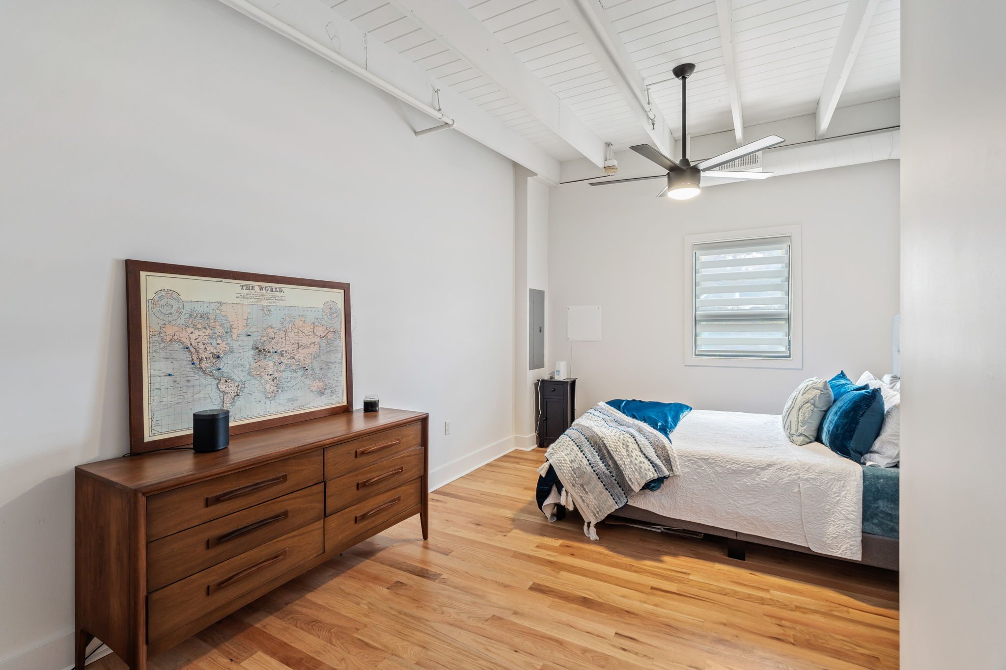 A minimalist bedroom with a wooden dresser, a world map above it, a bed with blue and white pillows, a throw blanket, a window with horizontal blinds, wood flooring, white walls, and a ceiling fan.