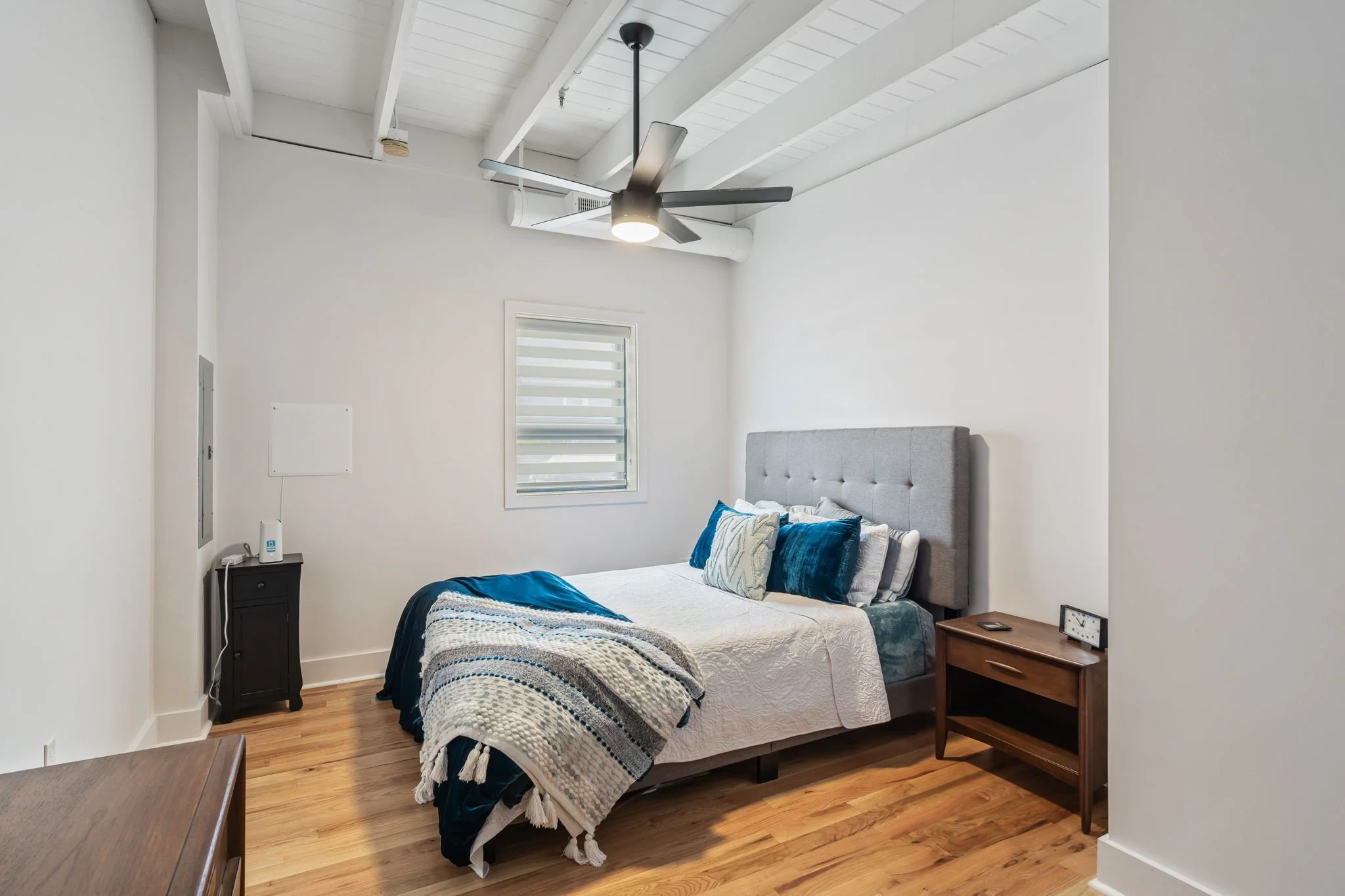 A modern bedroom with a gray upholstered bed, white bedding, and blue accent pillows, a wooden nightstand with a clock, a ceiling fan, and a small window with horizontal blinds, hardwood floors, and white walls.