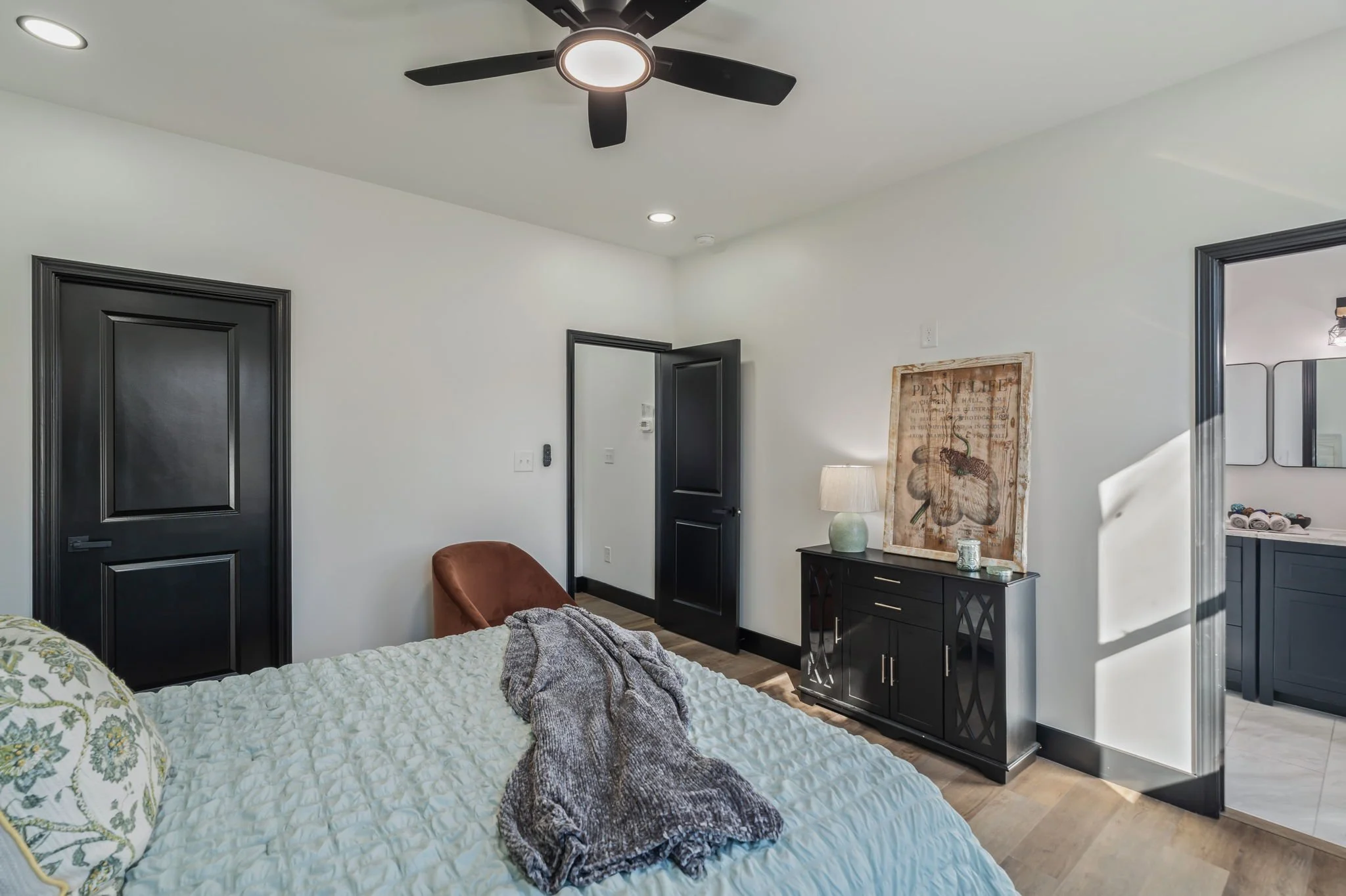 A modern bedroom with a bed, a black dresser with decorative items, a brown chair, and a doorway leading to a bathroom. Ceiling fan and recessed lighting visible.