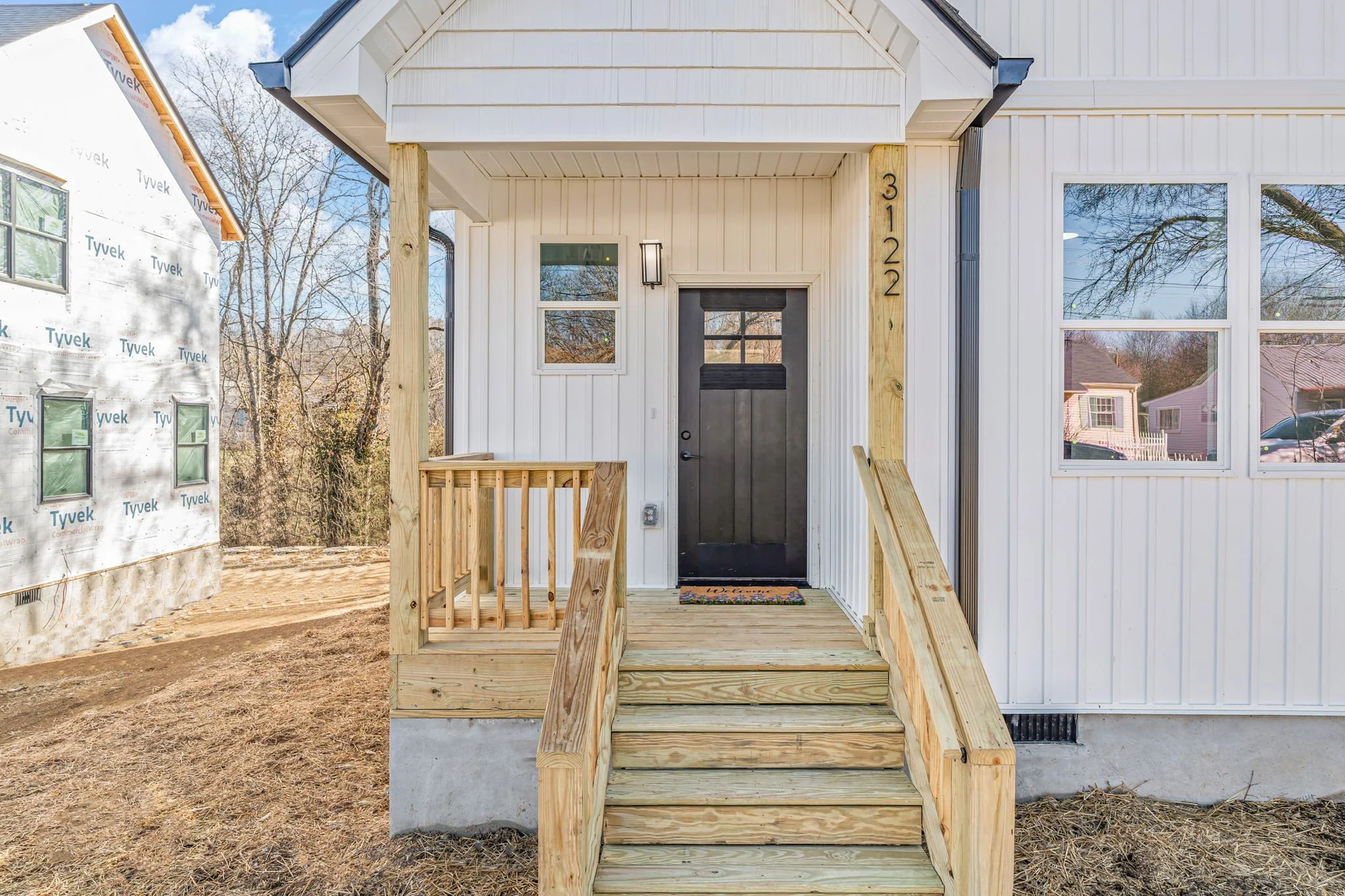 Front porch of a house under construction with a black front door, small window, and wooden stairs and railing. The house has white siding, and the house number 3122 is vertically displayed next to the door. To the left, there's a partially built str