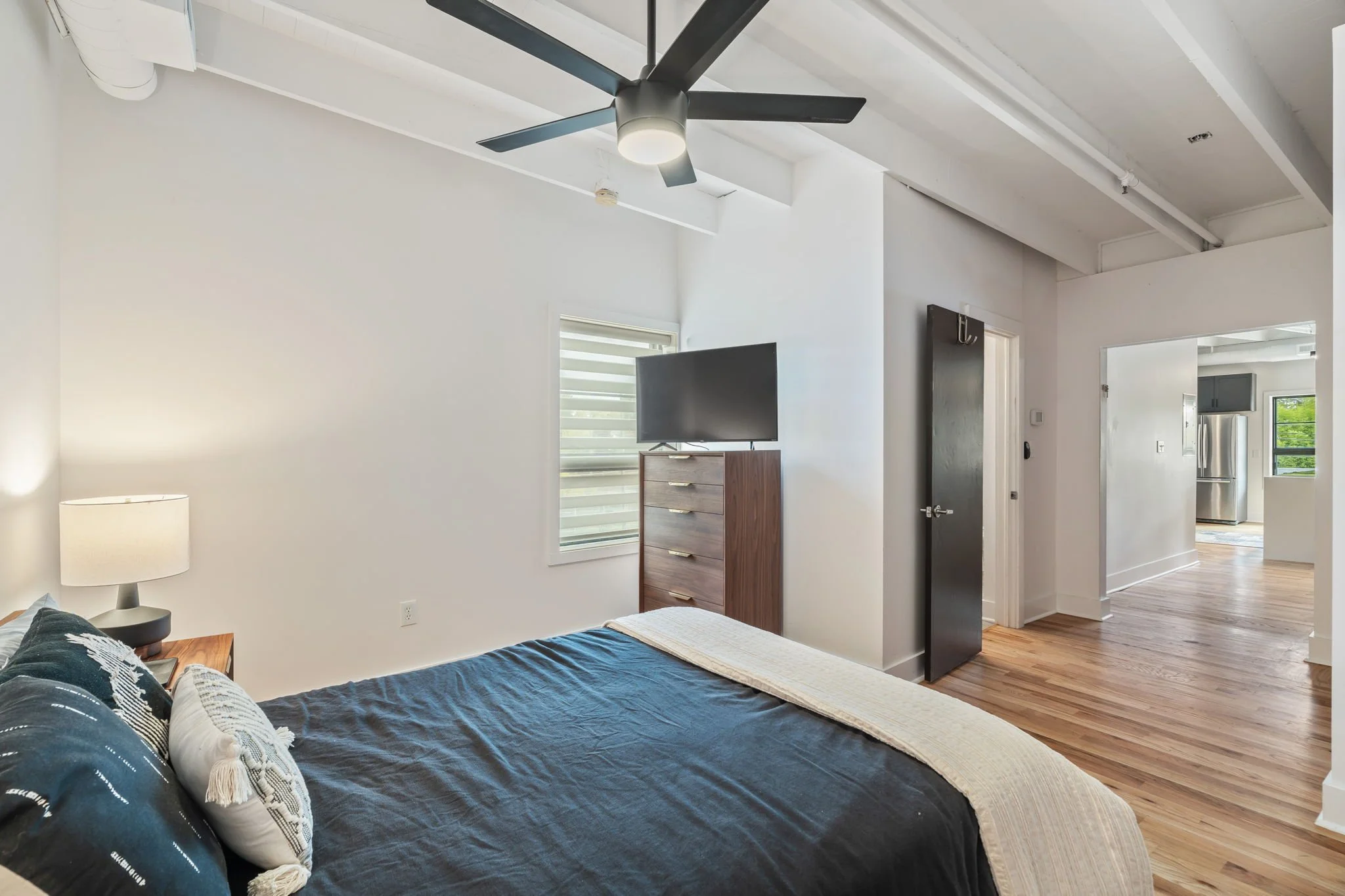 A bedroom with white walls, a ceiling fan, a bed with black bedding and pillows, a wooden nightstand with a lamp, a window with blinds, a dresser with a TV on top, and a door leading to a kitchen with stainless steel appliances and a window.