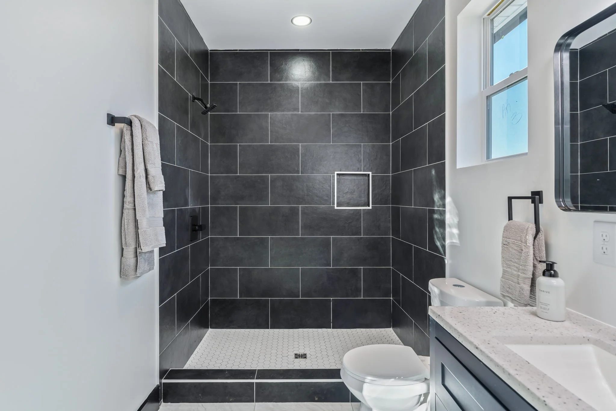 Modern bathroom with black tiled walk-in shower, a window, and a light-colored vanity with a soap dispenser.