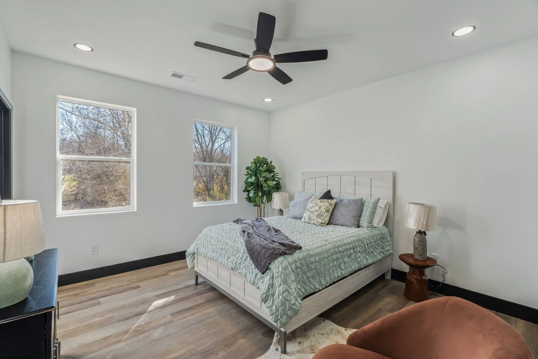 Clean bedroom with white walls, wooden flooring, double windows, a bed with a light-colored bedspread, and bedside lamps.
