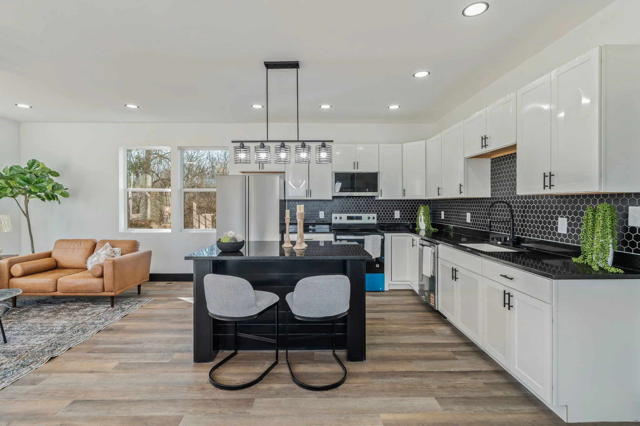 Modern open-concept kitchen with black countertops, white cabinets, black hexagonal backsplash, stainless steel appliances, and a dining island with two gray chairs. Adjacent to the kitchen is a living area with a tan leather sofa, decorative pillows