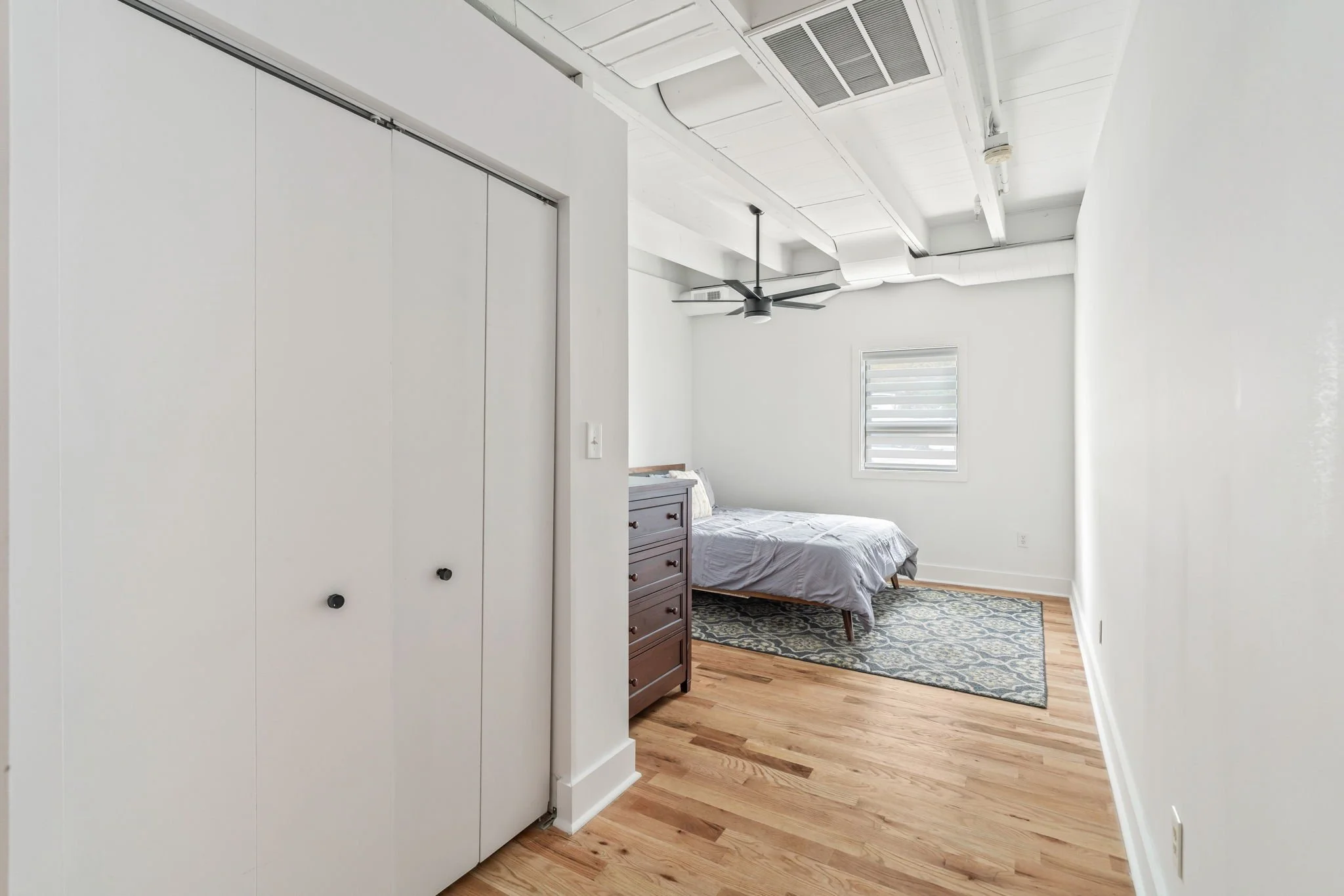 A minimalist bedroom with white walls, hardwood floors, a bed with white bedding, a small dresser, a window with blinds, and a ceiling fan.