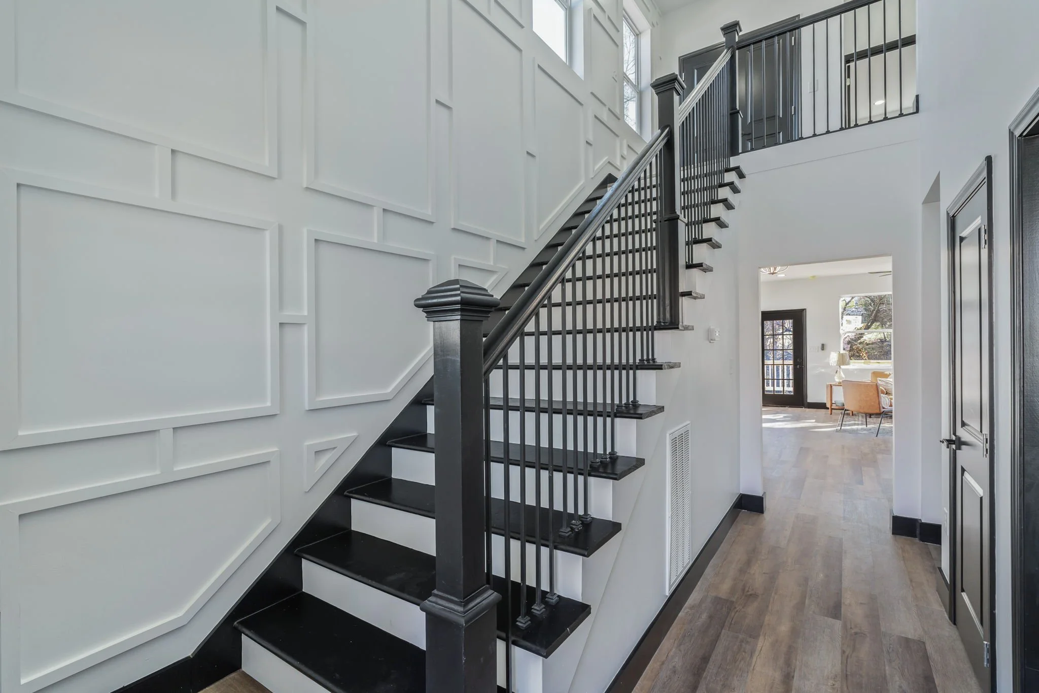 Interior view of a modern staircase with black handrails and steps, white wall with decorative molding, and a hallway leading to a room with large windows and seating.