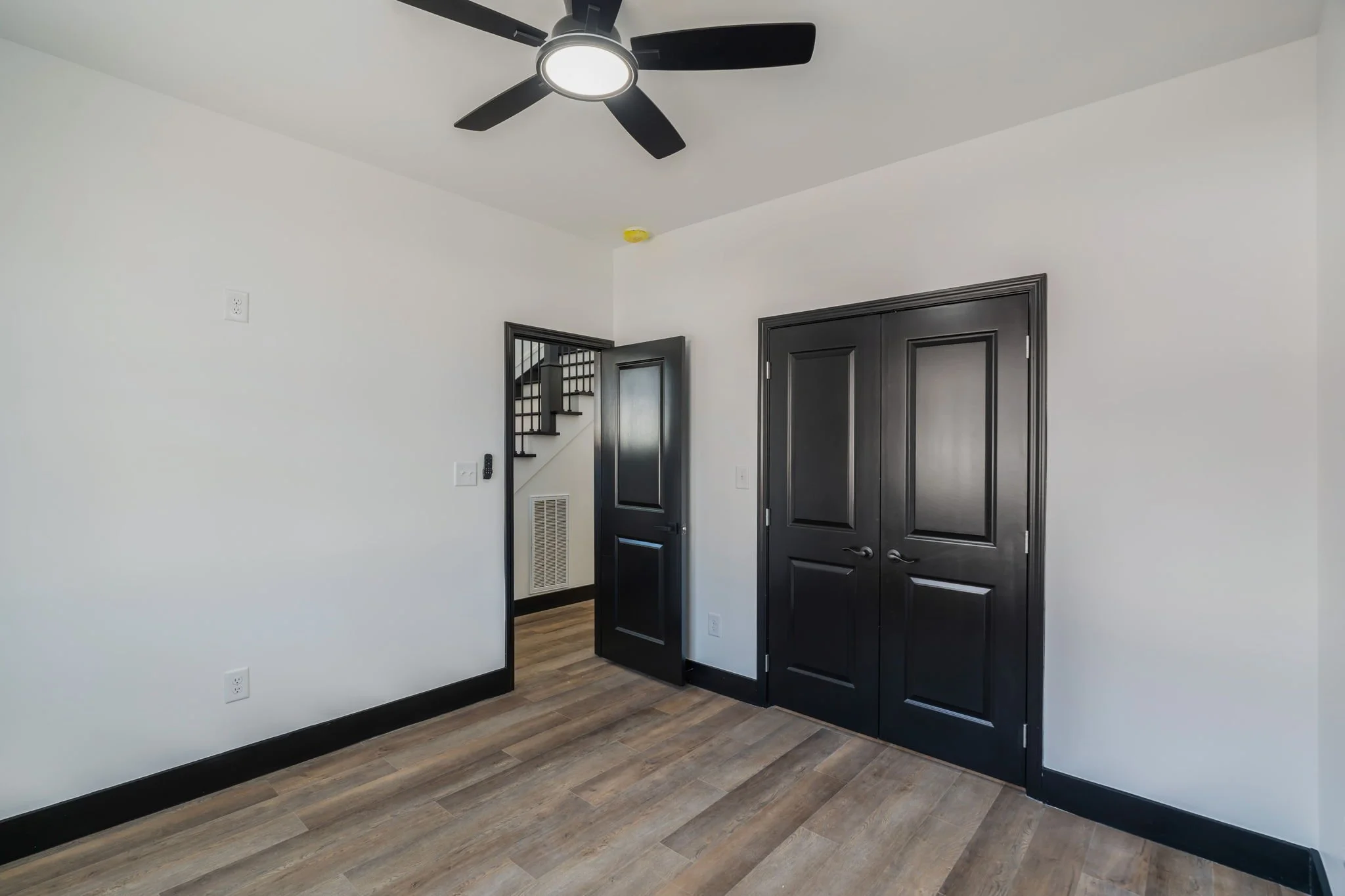 Empty room with white walls, a black ceiling fan with light, a black door, a black double closet door, wood-look flooring, and a staircase visible through an open door.