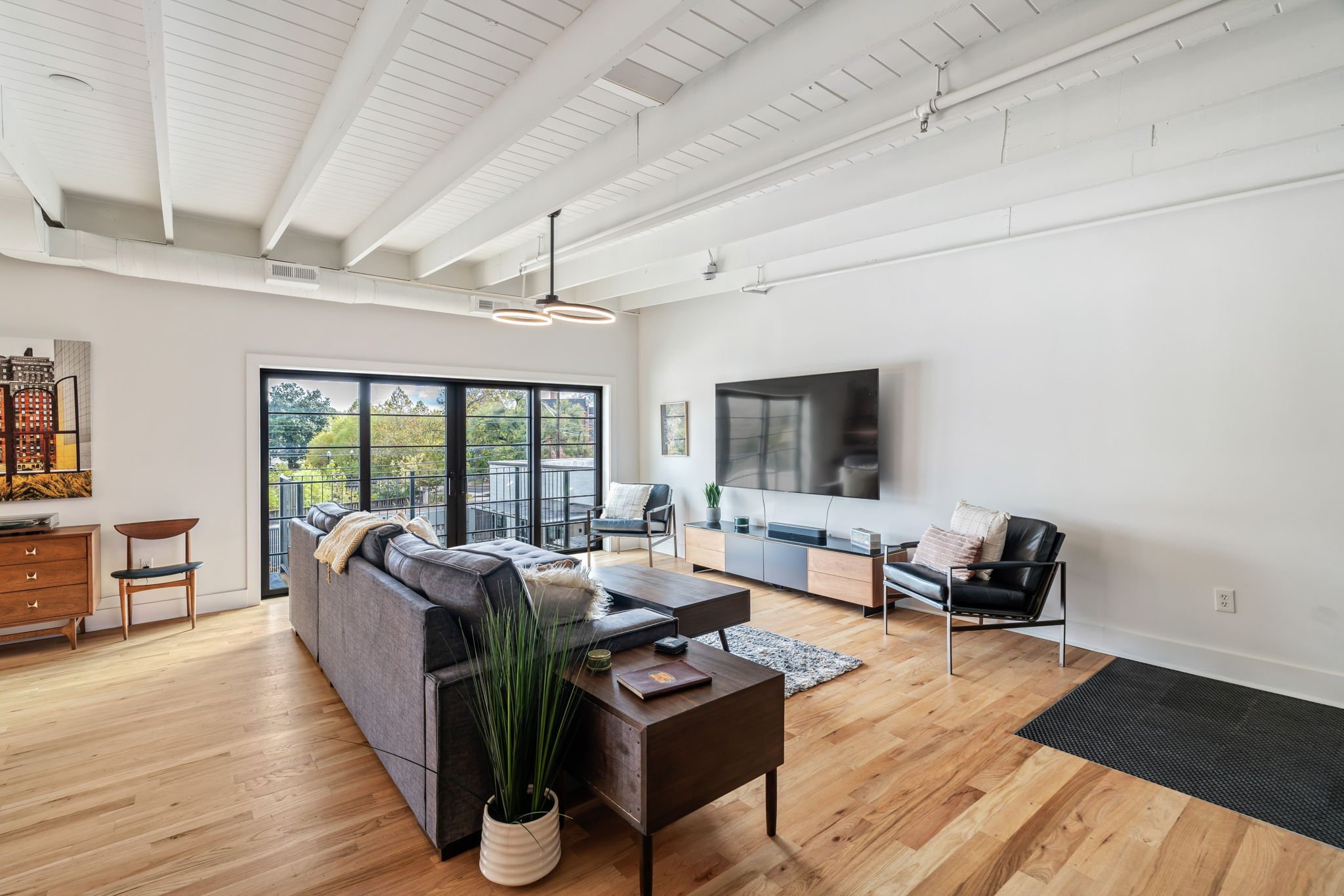 Modern living room with white ceiling beams, large black-framed windows, a mounted flat-screen TV, a gray sofa decorated with a blanket and pillows, black armchairs, and wooden furniture. Natural light floods the space, which has hardwood floors and 