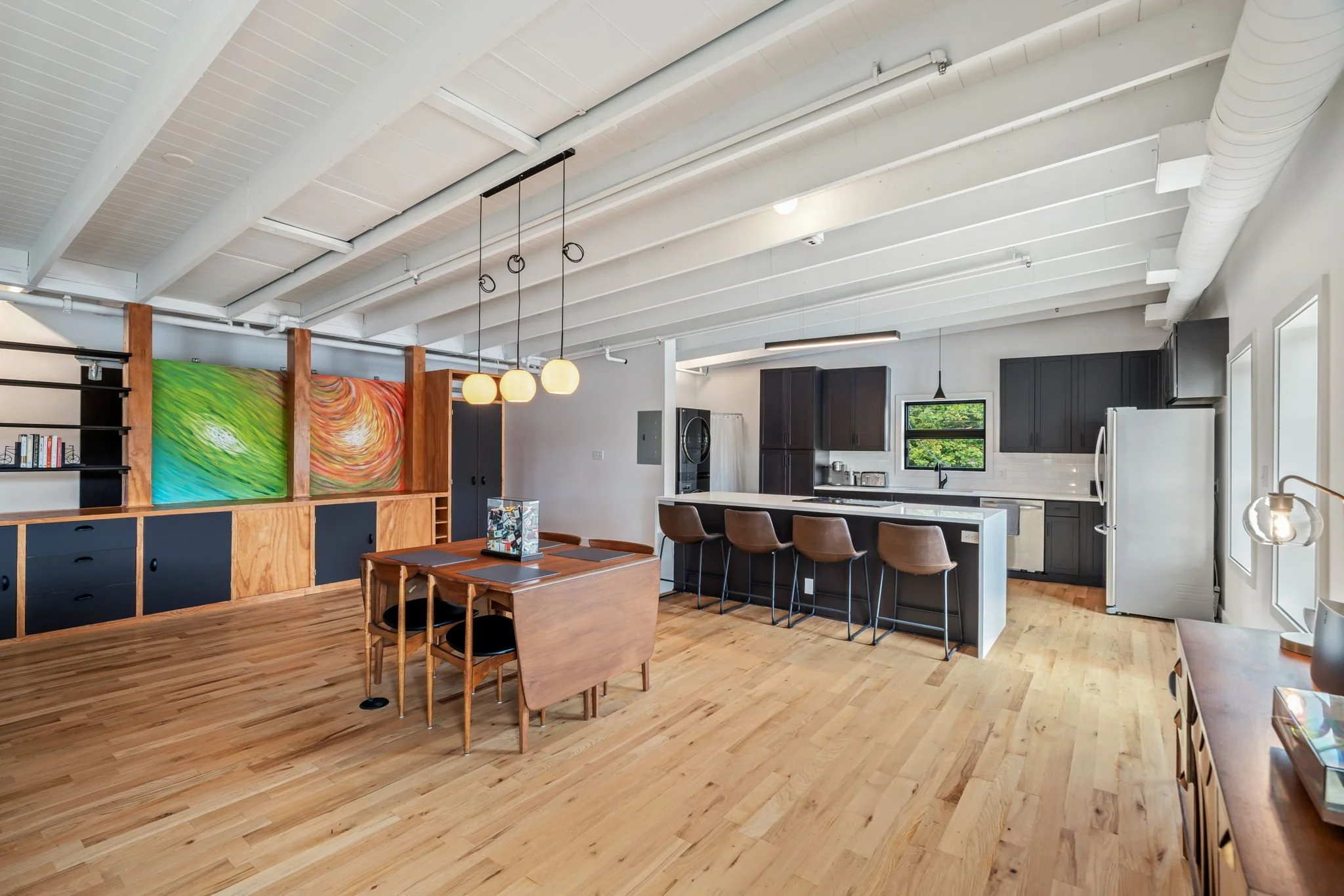 Open-plan kitchen and dining area with wooden flooring, black kitchen cabinets, a white countertop, barstools, and a colorful abstract art piece on a wooden partition.