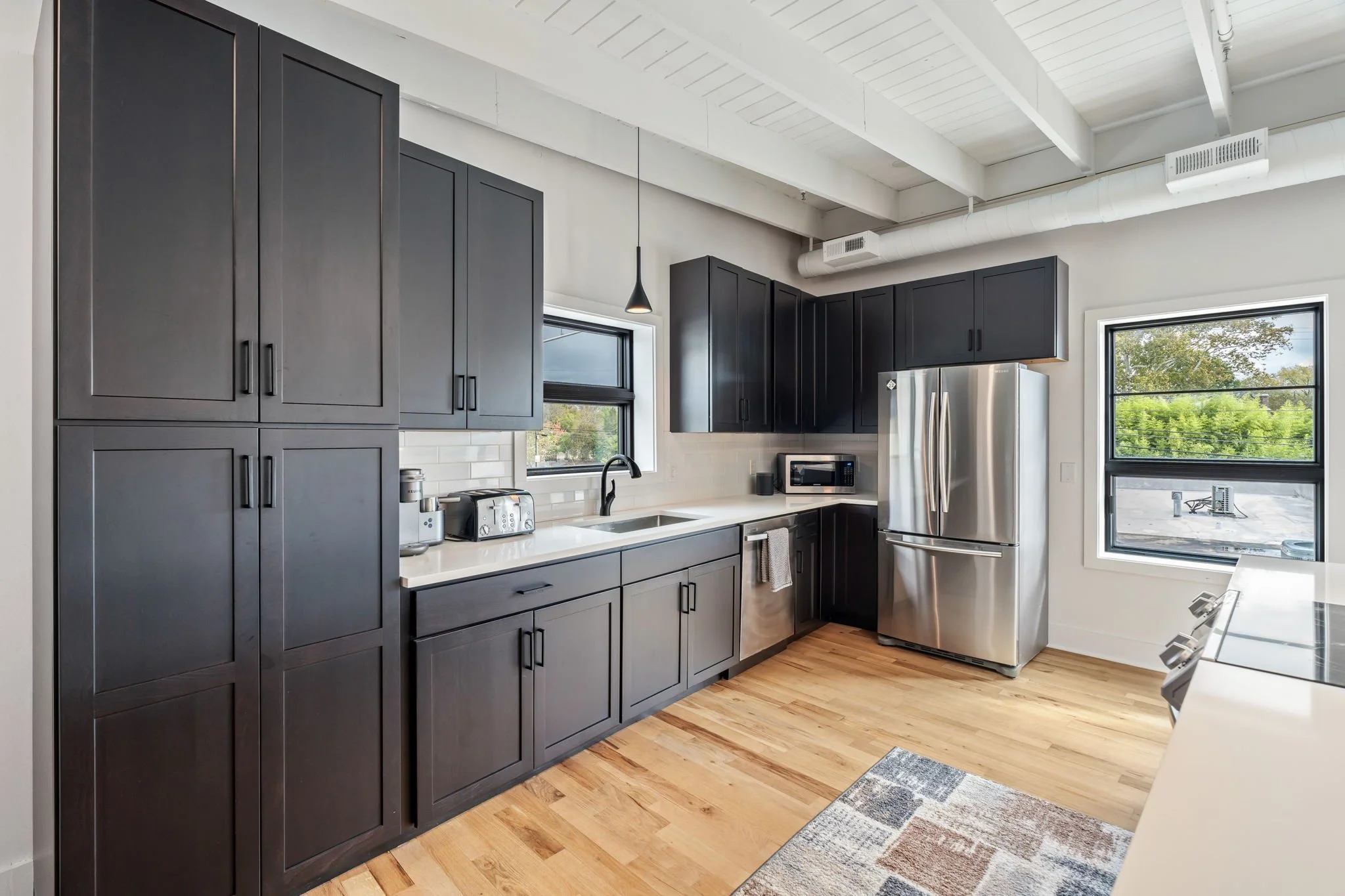 Modern kitchen with dark cabinets, stainless steel refrigerator, white countertops, hardwood floor, and two windows