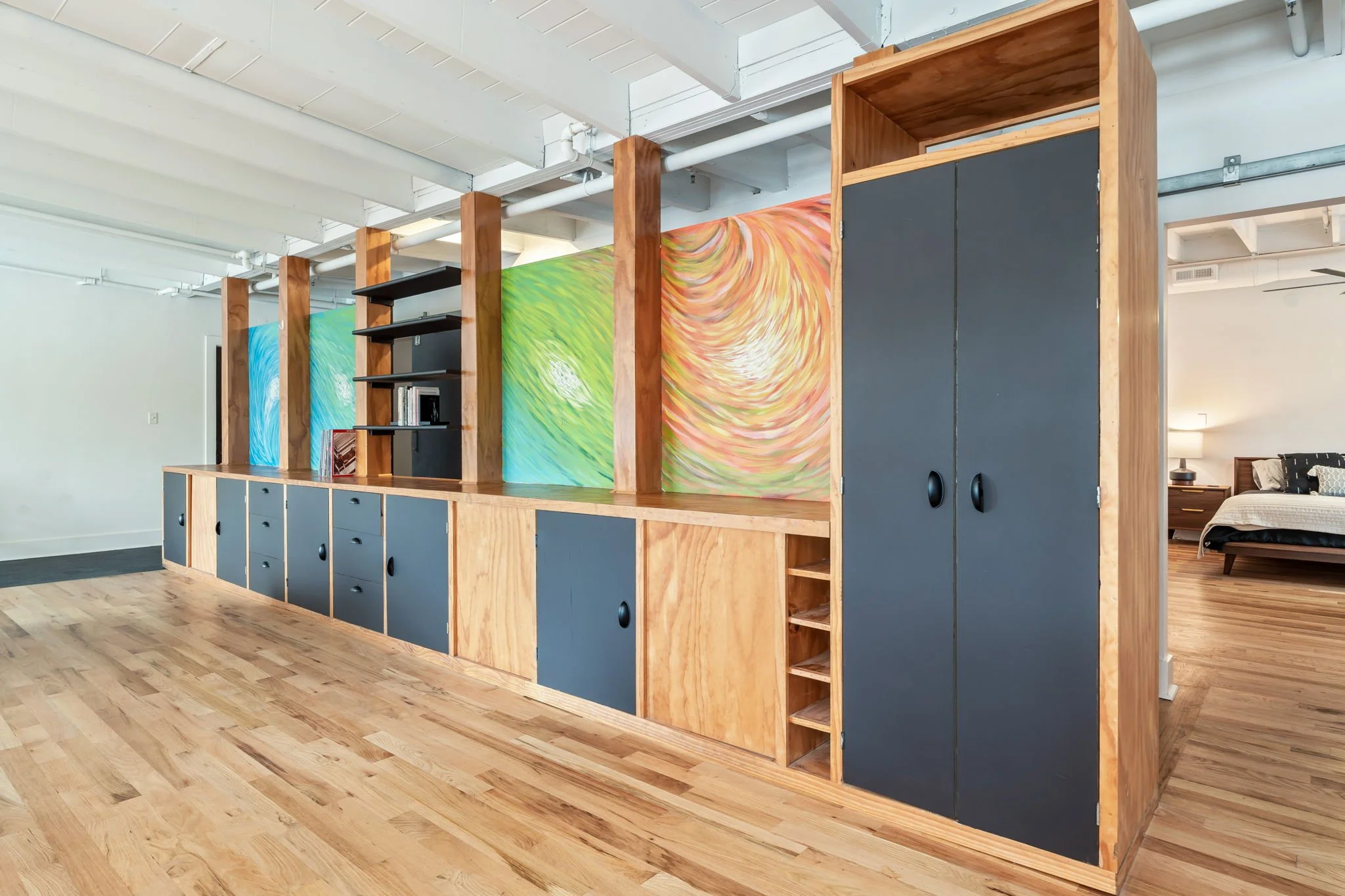 Modern wooden room divider with colorful swirl paintings, open shelves, and black cabinet doors, separating living space from bedroom.
