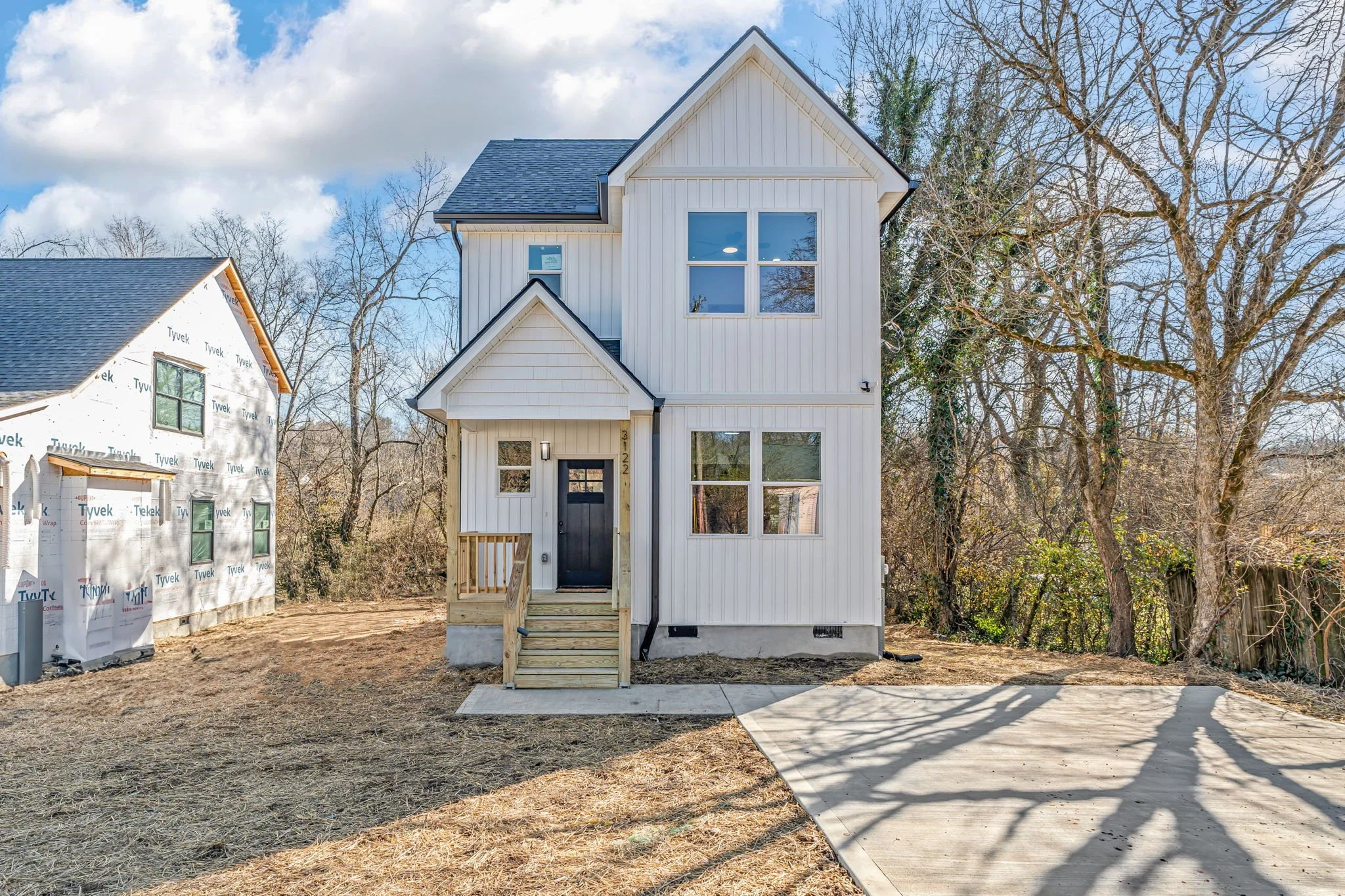 Newly built white two-story house with black door and front steps, next to a house under construction, on a partly cloudy day with trees around