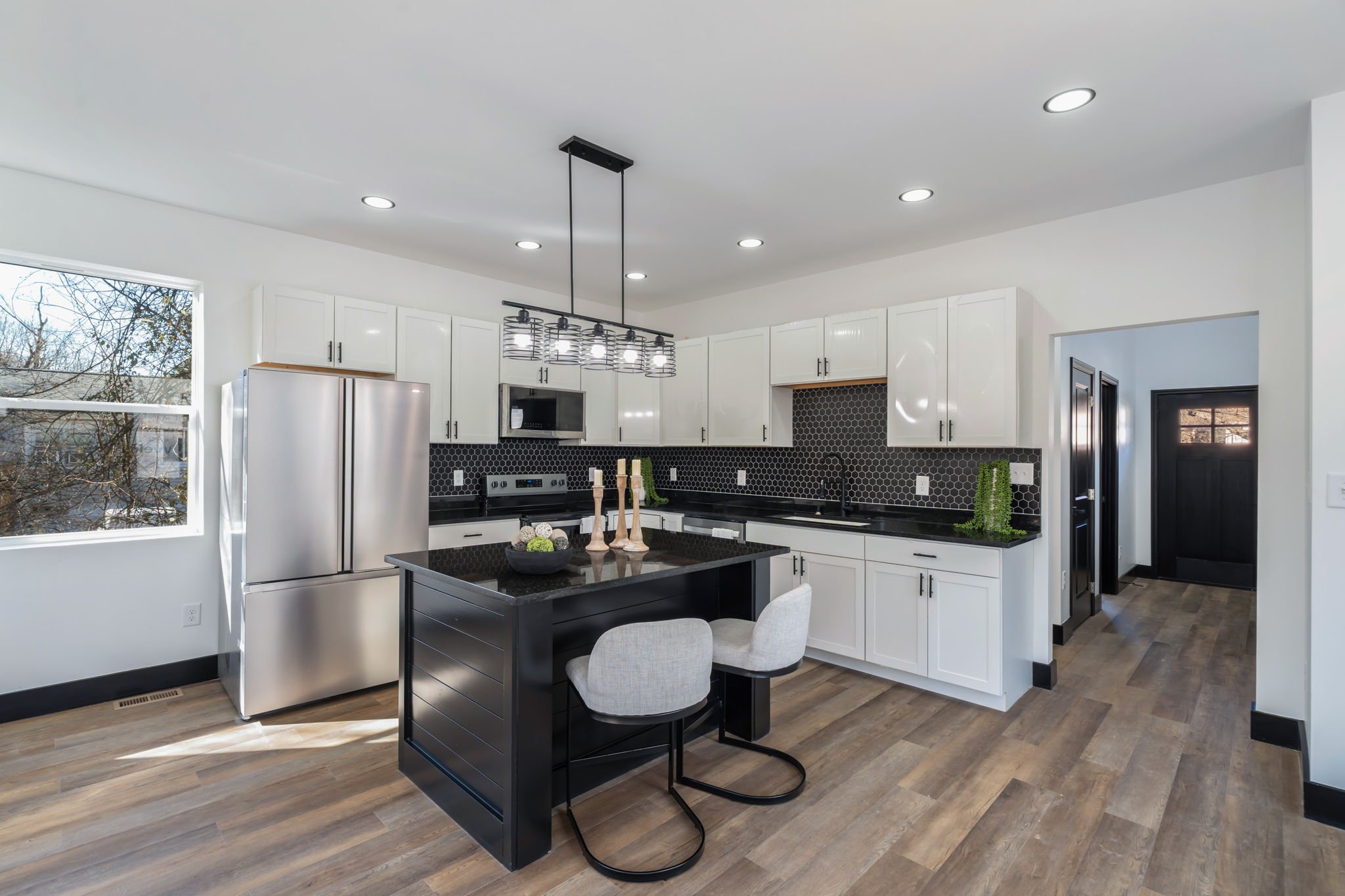 Modern kitchen with white cabinets, black countertops, stainless steel appliances, and a black island with two beige chairs. Black hexagonal backsplash, wooden flooring, and modern light fixture. Two large windows showing trees outside.
