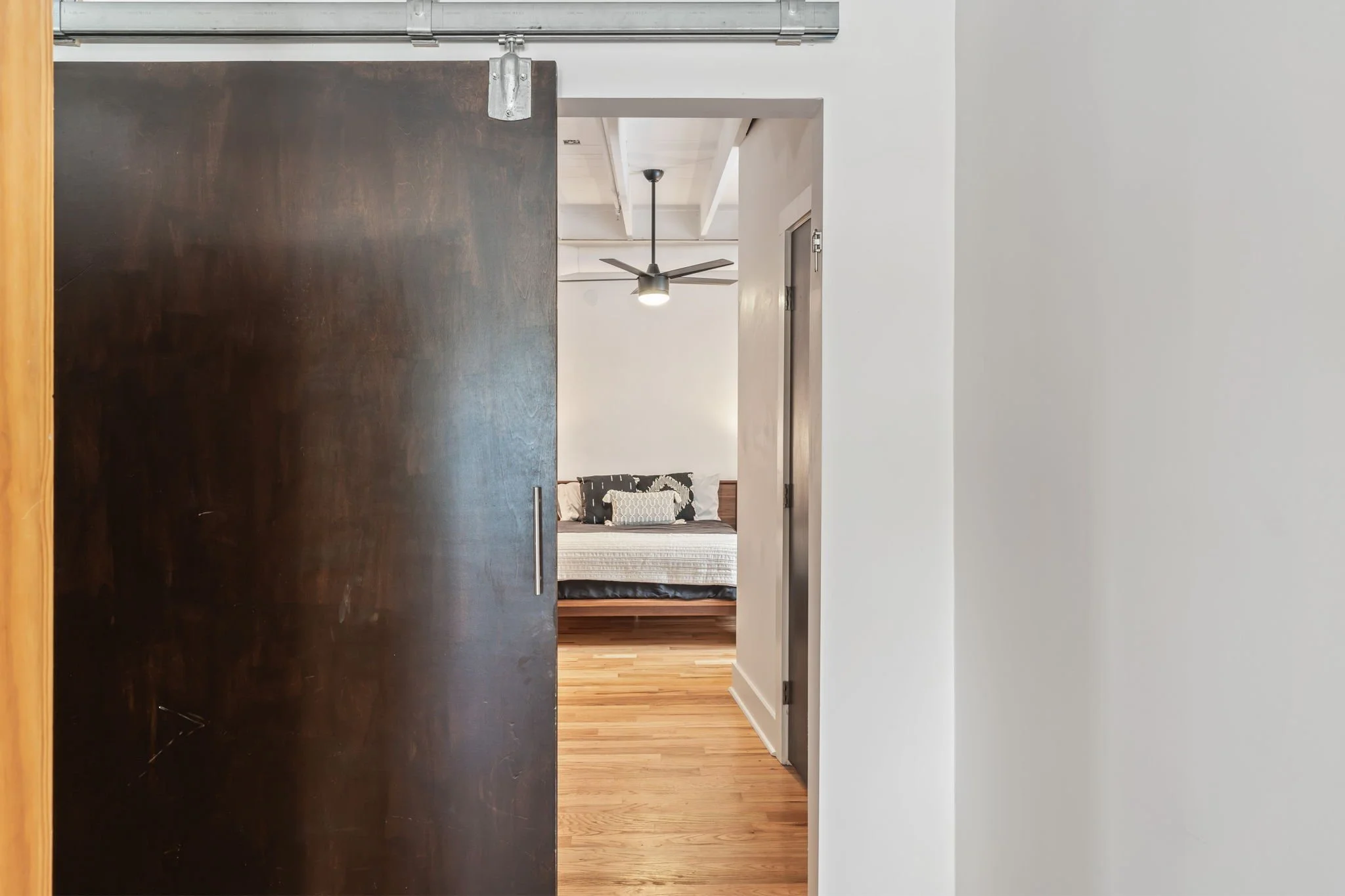View of a bedroom through a partially open dark wooden sliding door, with a bed and ceiling fan visible in the background.