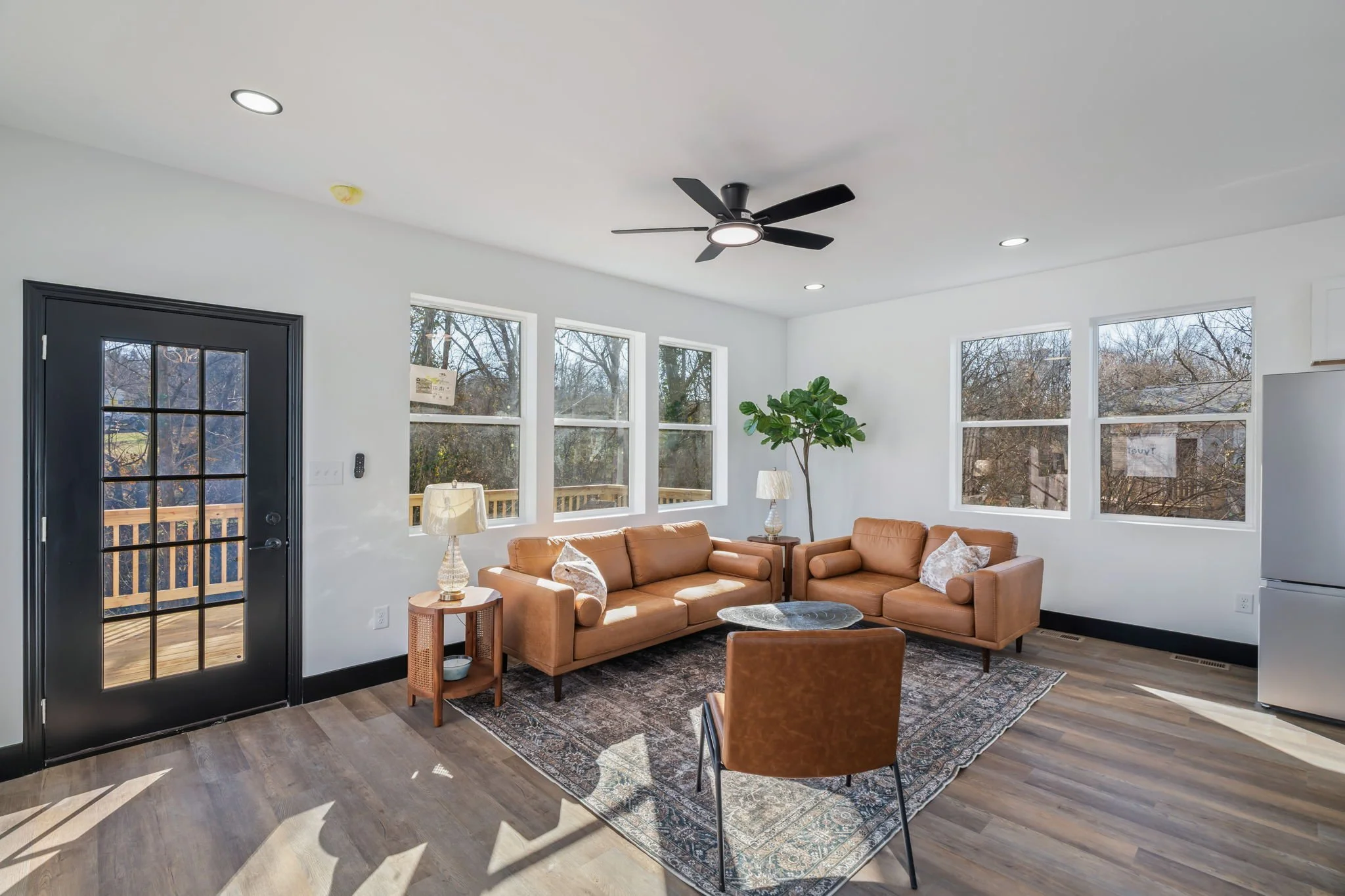 Living room with tan leather sofas, a patterned rug, a black ceiling fan, and large windows showing outdoor trees and a deck.