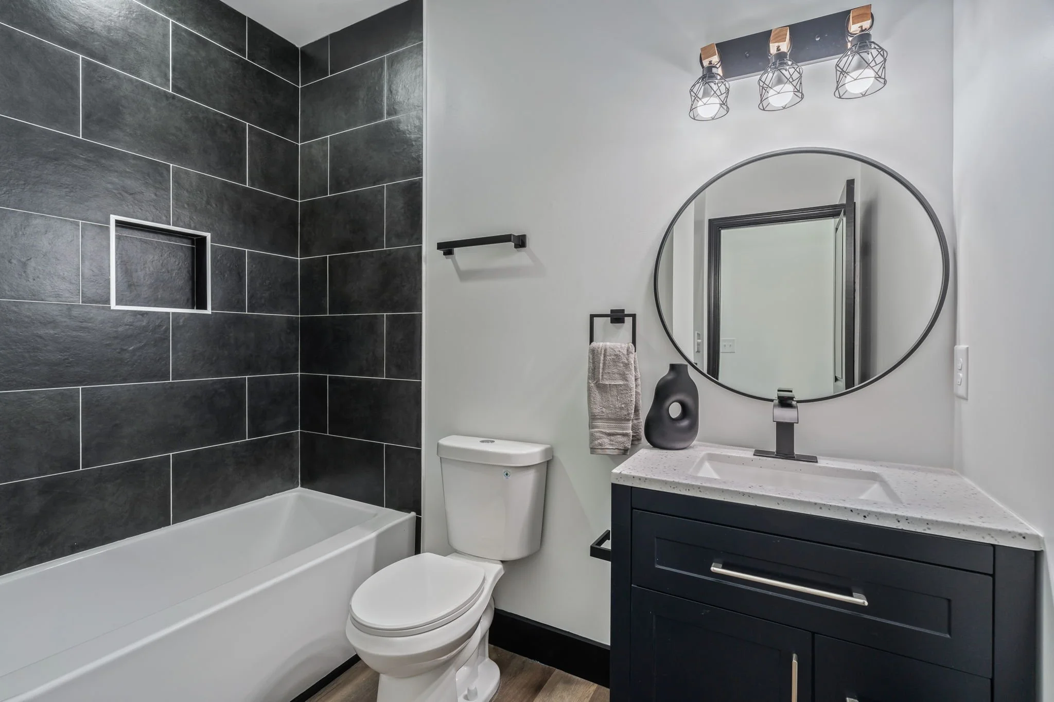 Modern bathroom with black tiled shower, white toilet, and black vanity with speckled countertop, oval mirror, and black light fixtures.
