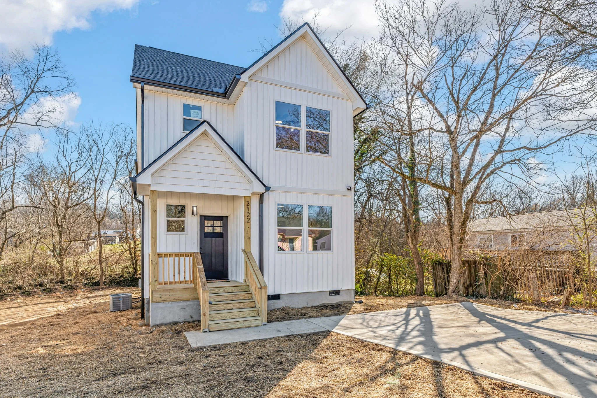 Newly built, white multi-story house with black door, small porch, wooden stairs, and concrete driveway, surrounded by leafless trees on a sunny day.