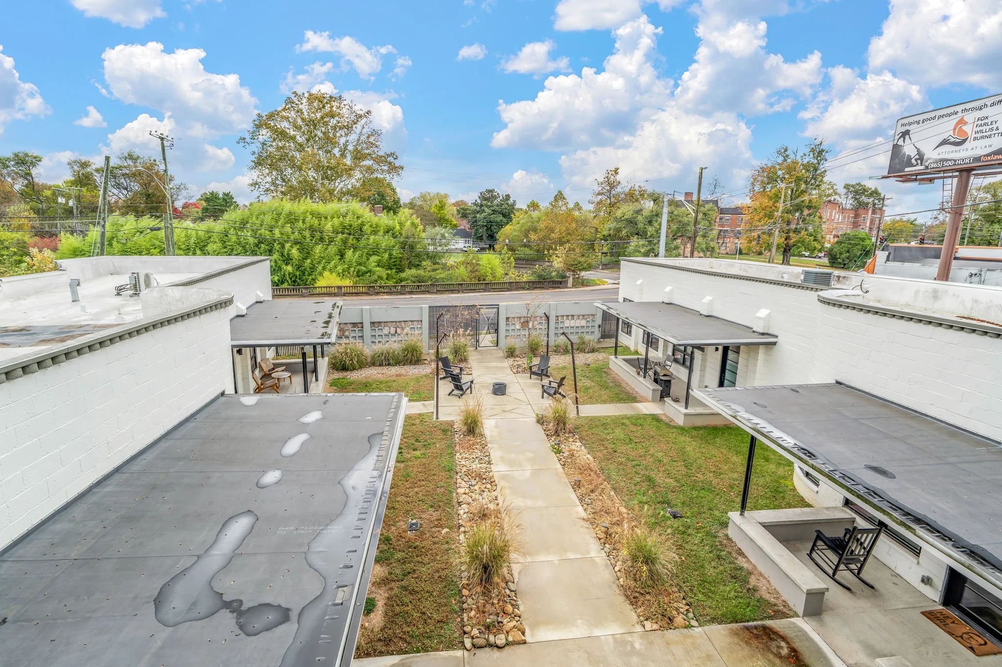View of a small outdoor courtyard with a concrete pathway, surrounded by white brick buildings with flat roofs, some with awnings and seating areas. There are four chairs arranged around a fire pit in the center of the courtyard. The sky is partly cl