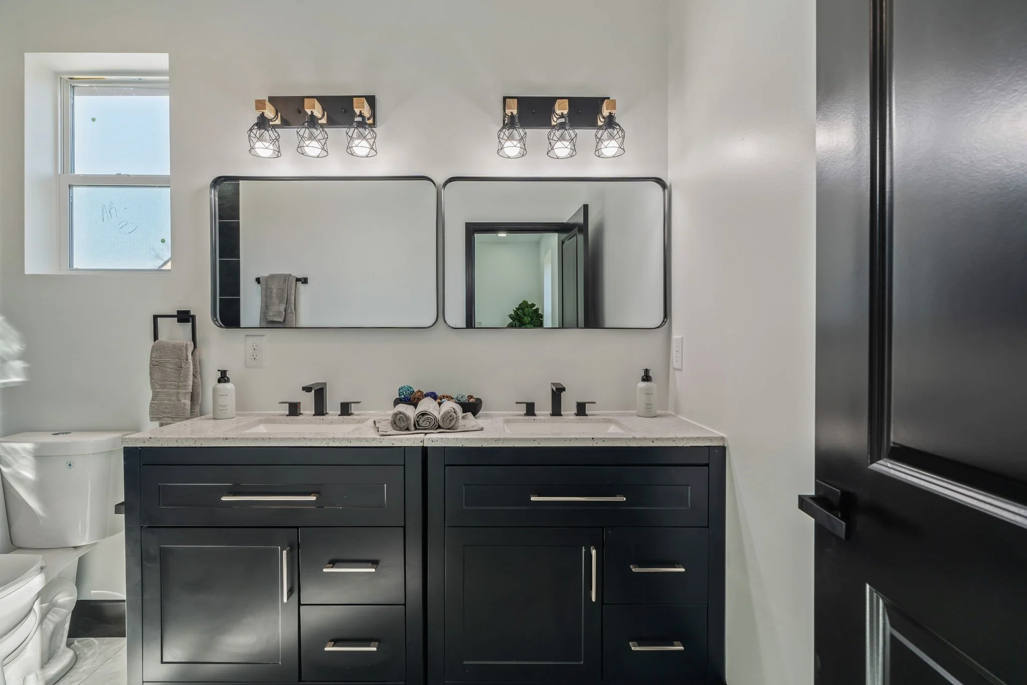 Modern bathroom with dual vanity, black cabinets, white countertop, two sinks, two rectangular mirrors, black faucets, wall-mounted light fixtures with multiple bulbs, window with frosted glass, towel hanging on a black towel bar, soap dispensers, an