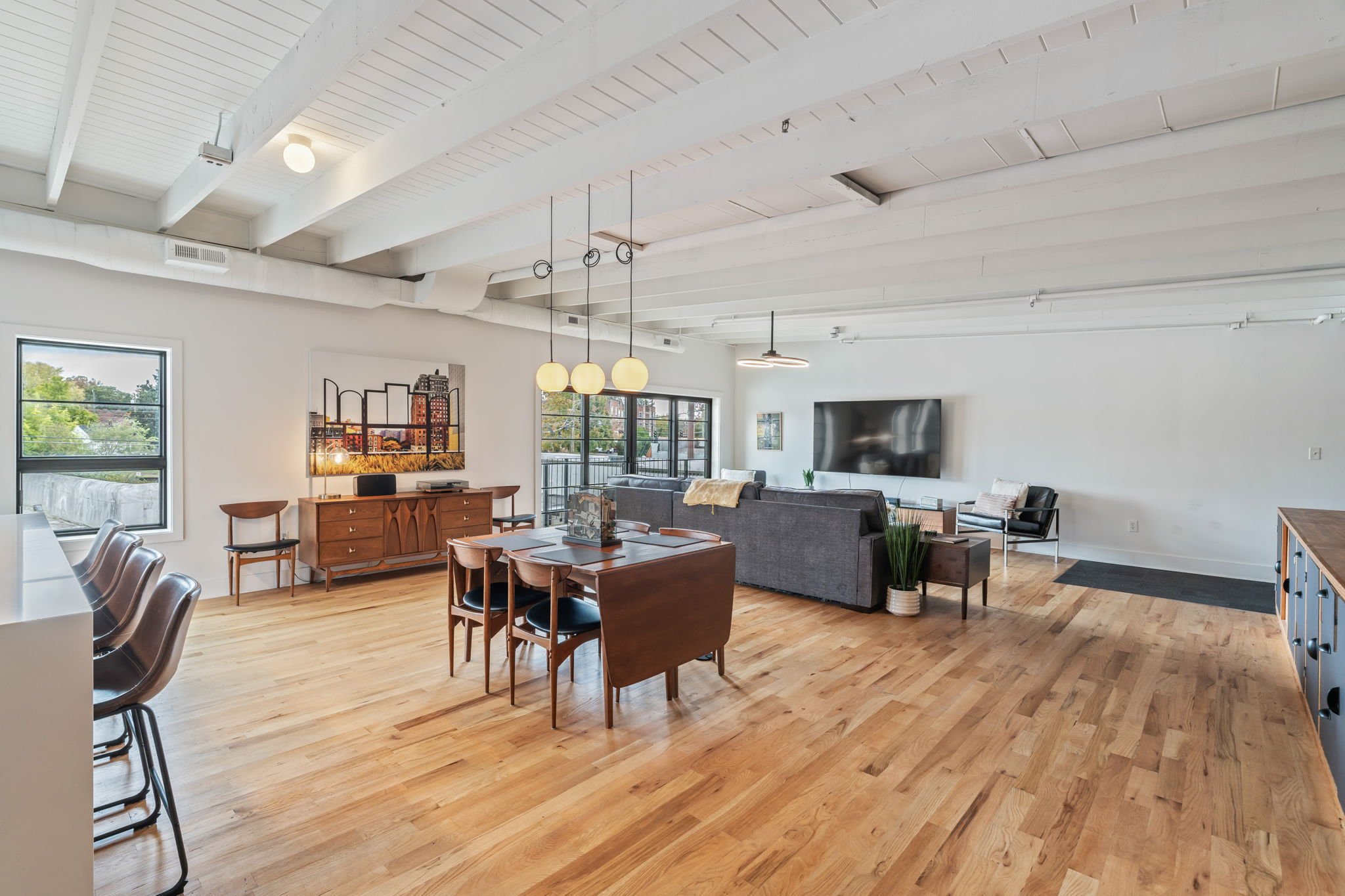 Bright open living and dining area with hardwood floors, white exposed ceiling beams, a gray sectional sofa, a tabletop dining set, a wooden sideboard, and a large flat-screen TV on the wall.