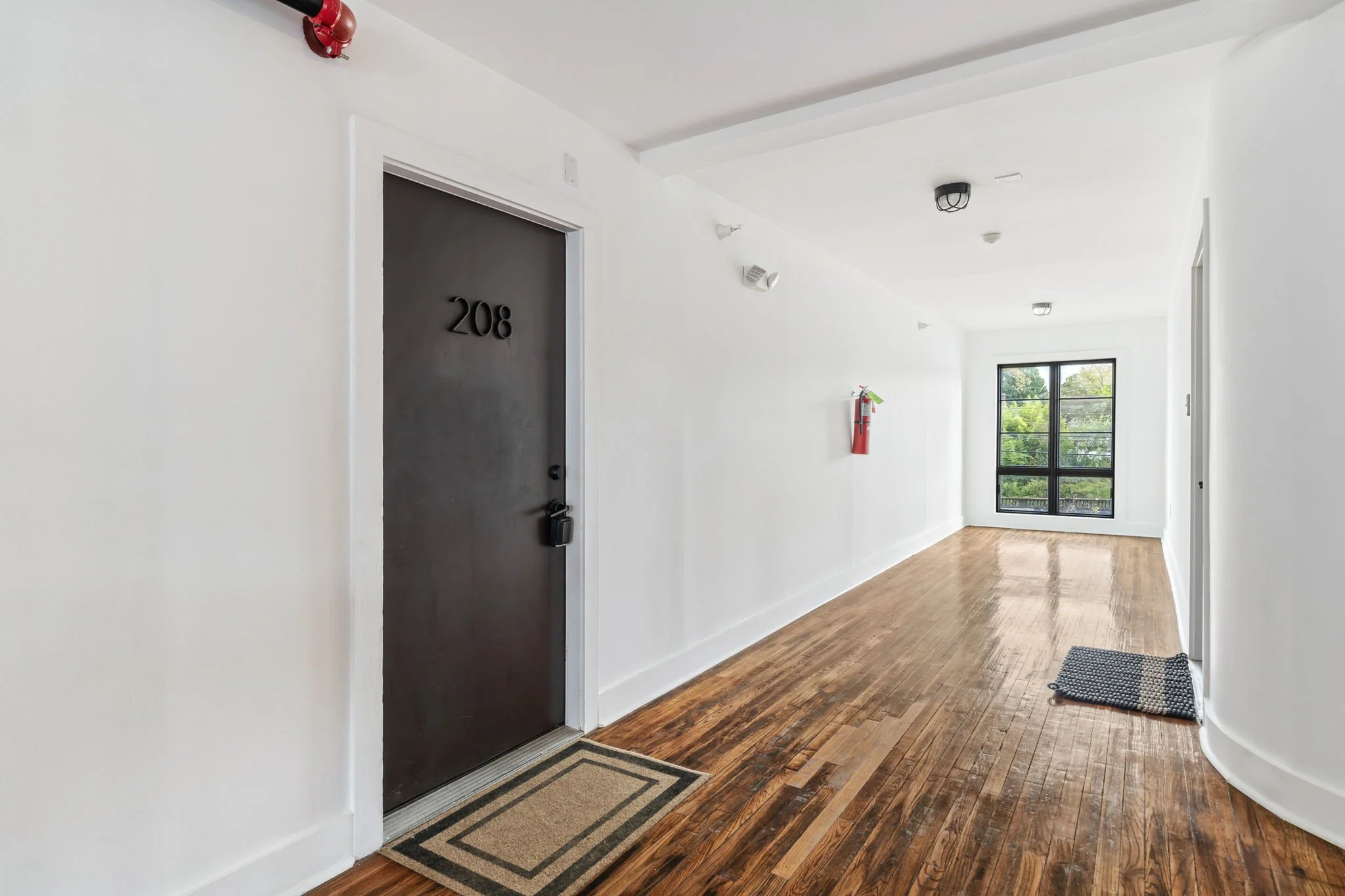 Empty hallway with hardwood floor, white walls, and a large window at the end. A black door labeled 208 with a rug in front, and a fire extinguisher mounted on the wall.