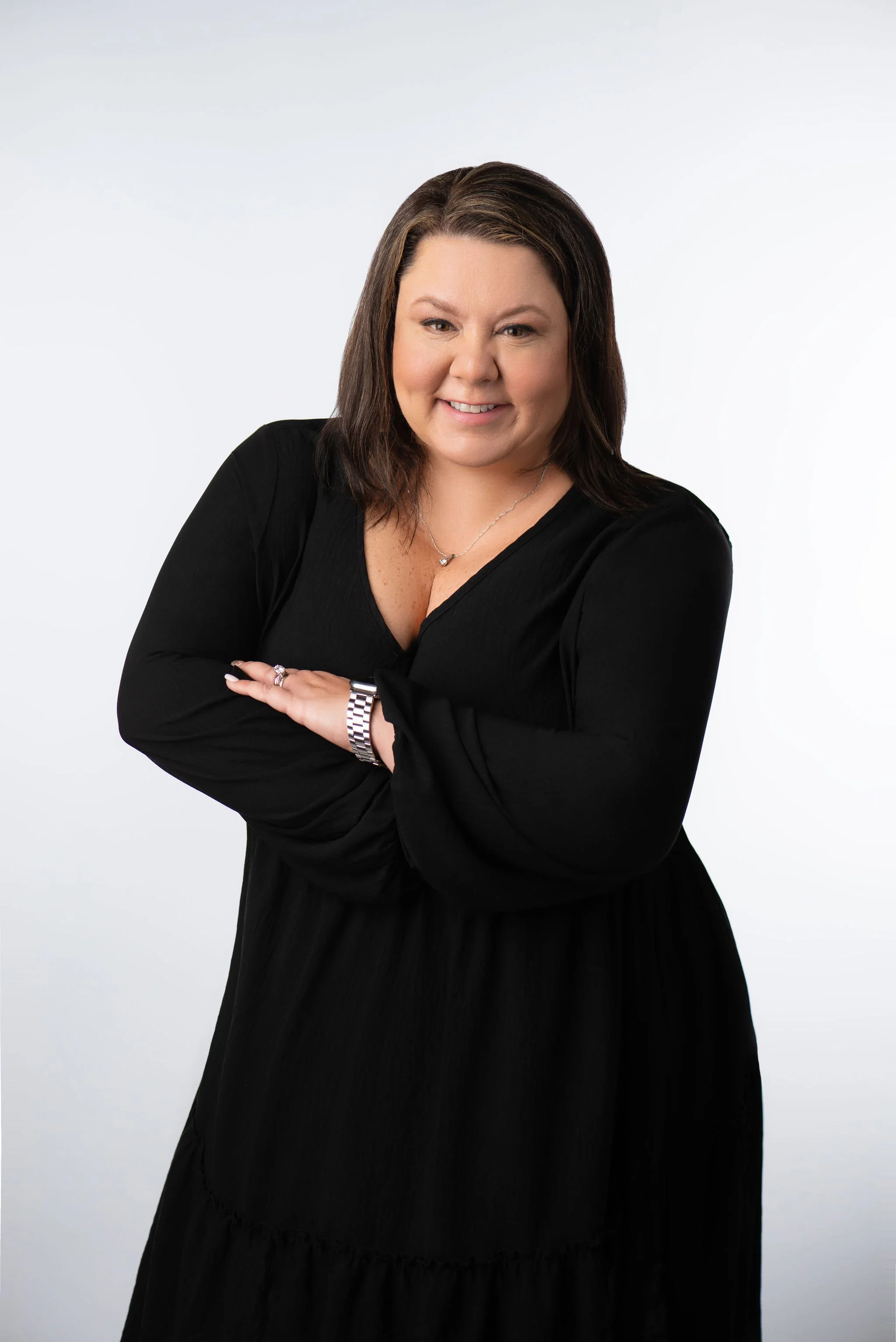 Woman in black dress with arms crossed, smiling, on a light background.