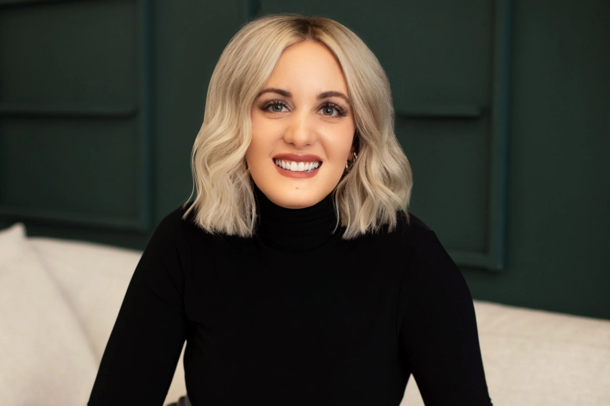 Person with blonde hair wearing a black turtleneck, smiling in front of a dark green background.