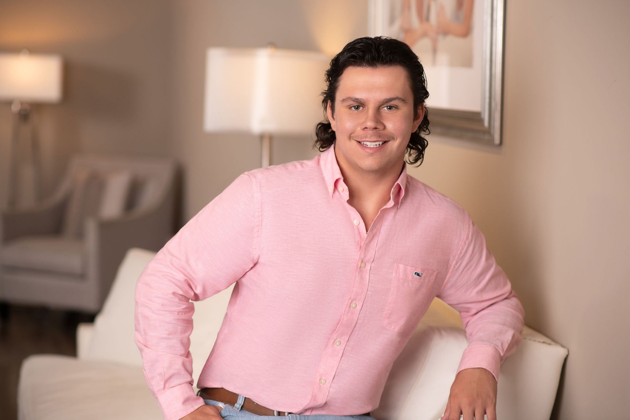 Smiling person in a pink shirt seated on a white couch, with a lamp and framed artwork in the background.
