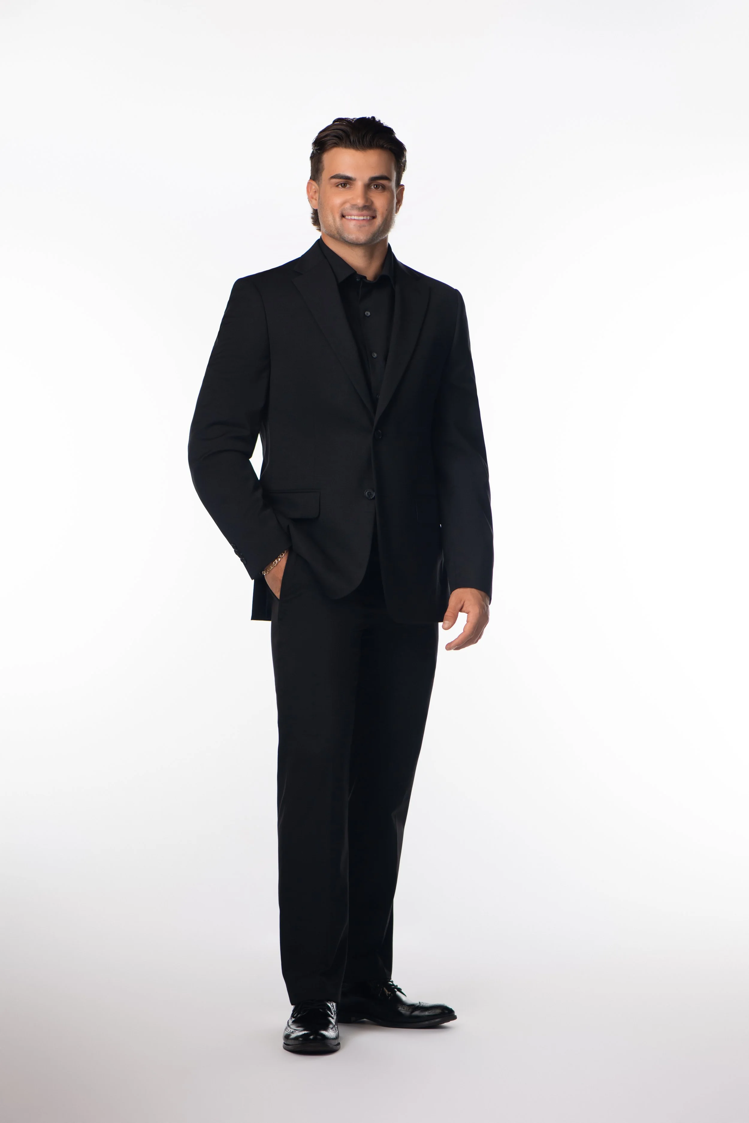 Man in a black suit smiling with hands in pockets against white background.