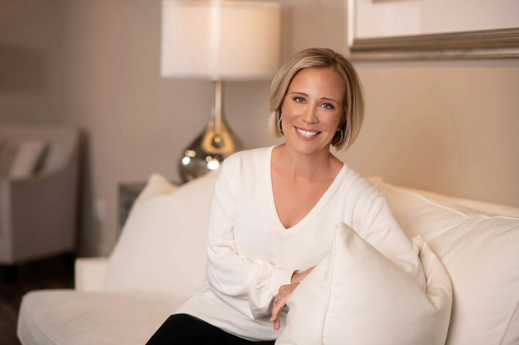 Person sitting on a couch in a cozy living room, smiling, with a lamp and cushions.