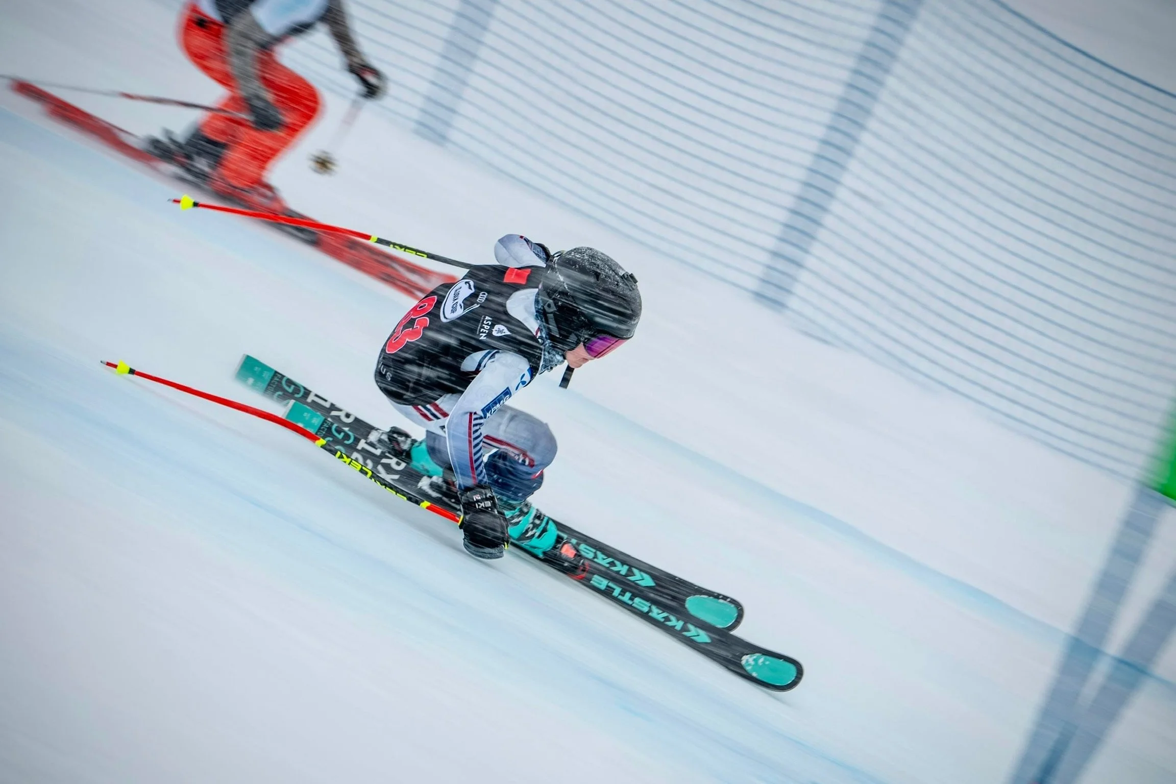 A skier racing downhill on a snowy slope, wearing a black helmet, black and white racing suit, and blue skis, with a safety net and snow-covered ground in the background.