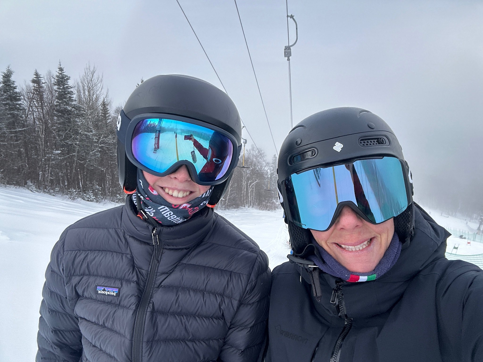 Two skiers wearing helmets and goggles smiling at the snow-covered ski resort, with ski lifts and trees in the background.