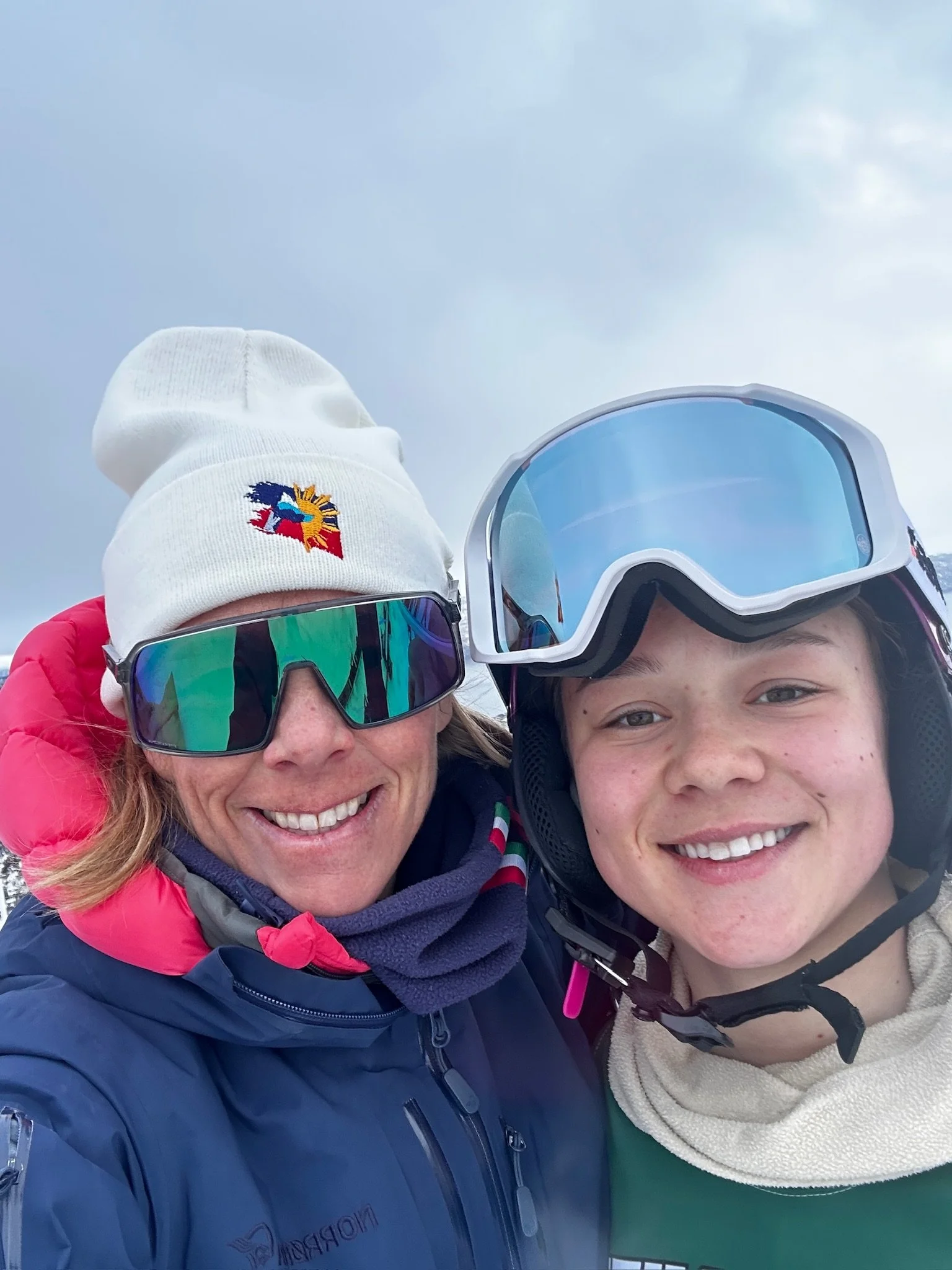 Two women in ski gear, both wearing helmets and goggles, smiling for a selfie outdoors with a cloudy sky in the background.