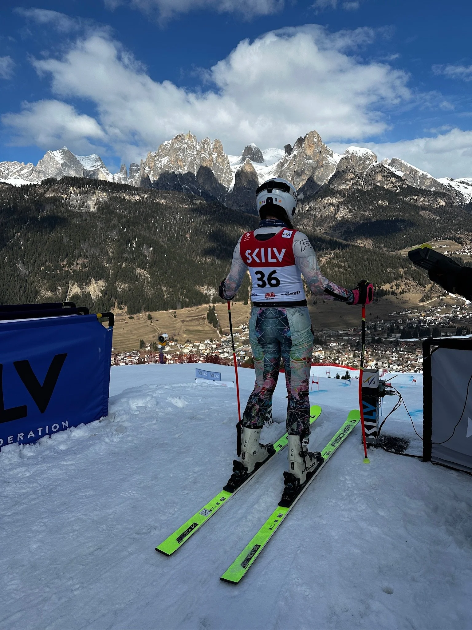 A skier wearing a red and white racing suit with the name 'SKILV' and the number 36 on the back, standing on yellow racing skis on a snowy slope, with a mountain range in the background and a blue sky with scattered clouds.