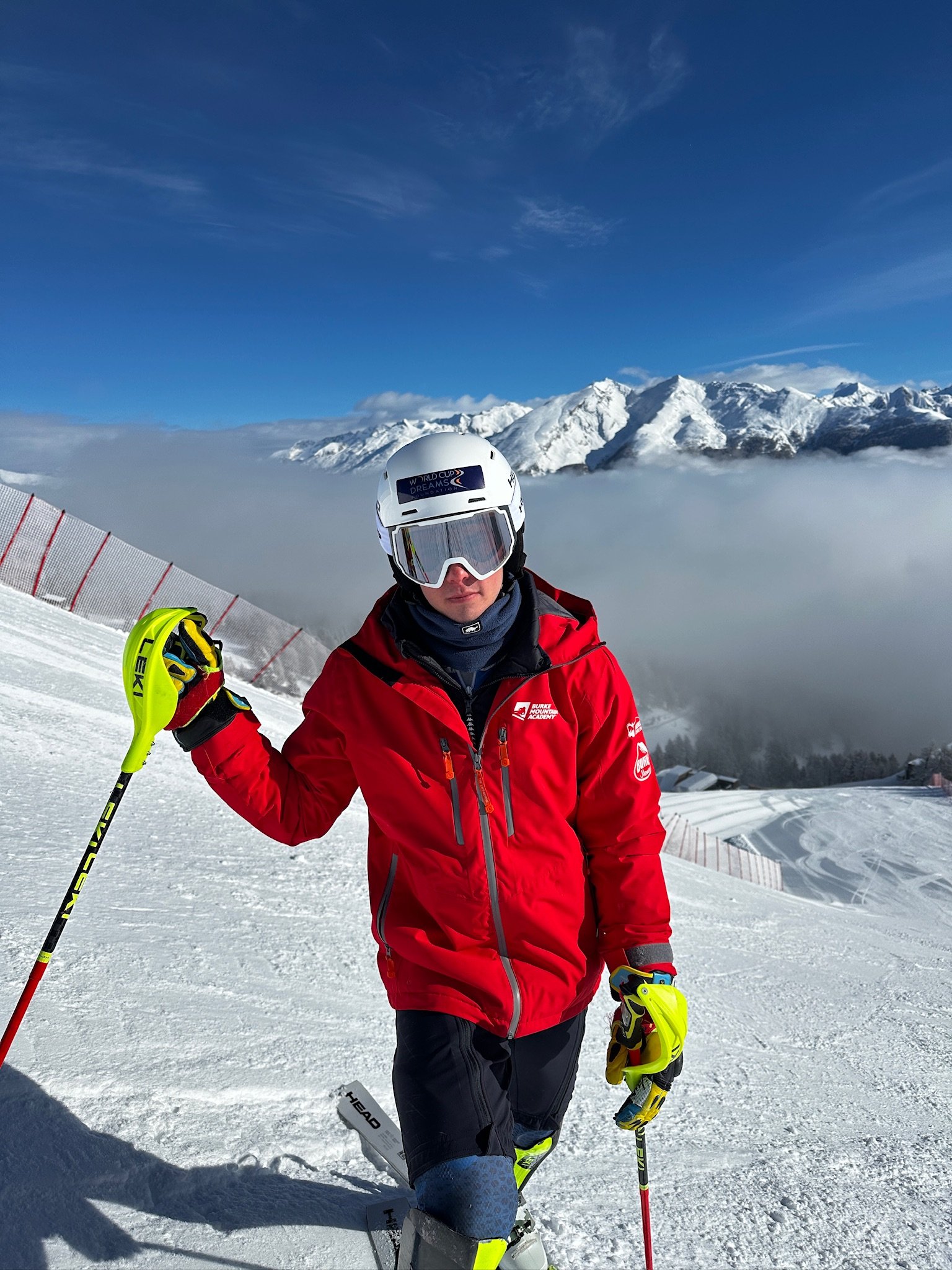 A skier dressed in a red jacket, black pants, and a white helmet is standing on a snowy slope with snow-covered mountains in the background under a blue sky.