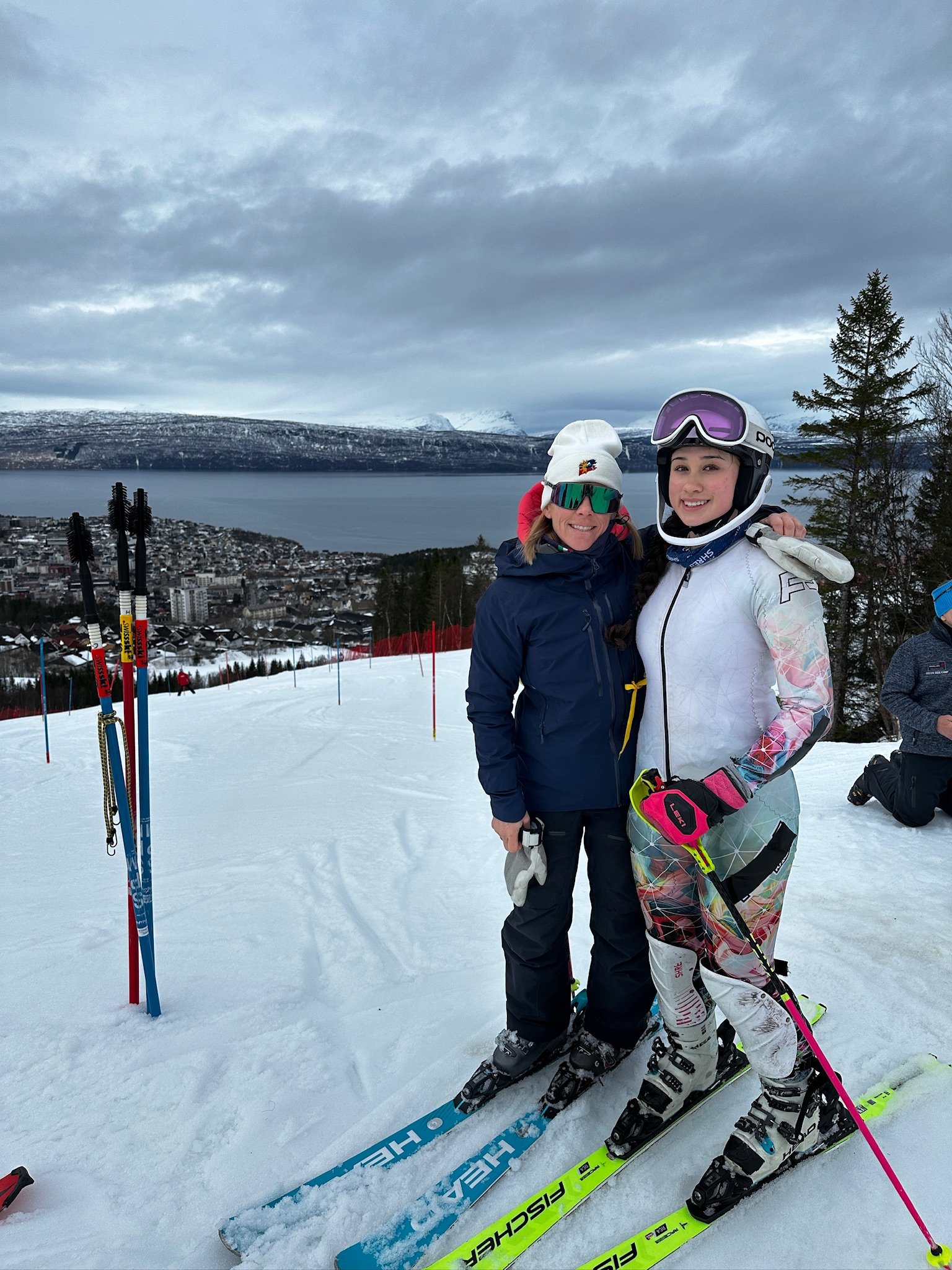 Two women dressed in winter ski gear smiling and posing on a snowy mountain. Snow-covered town, lake, and mountains in the background with overcast sky.