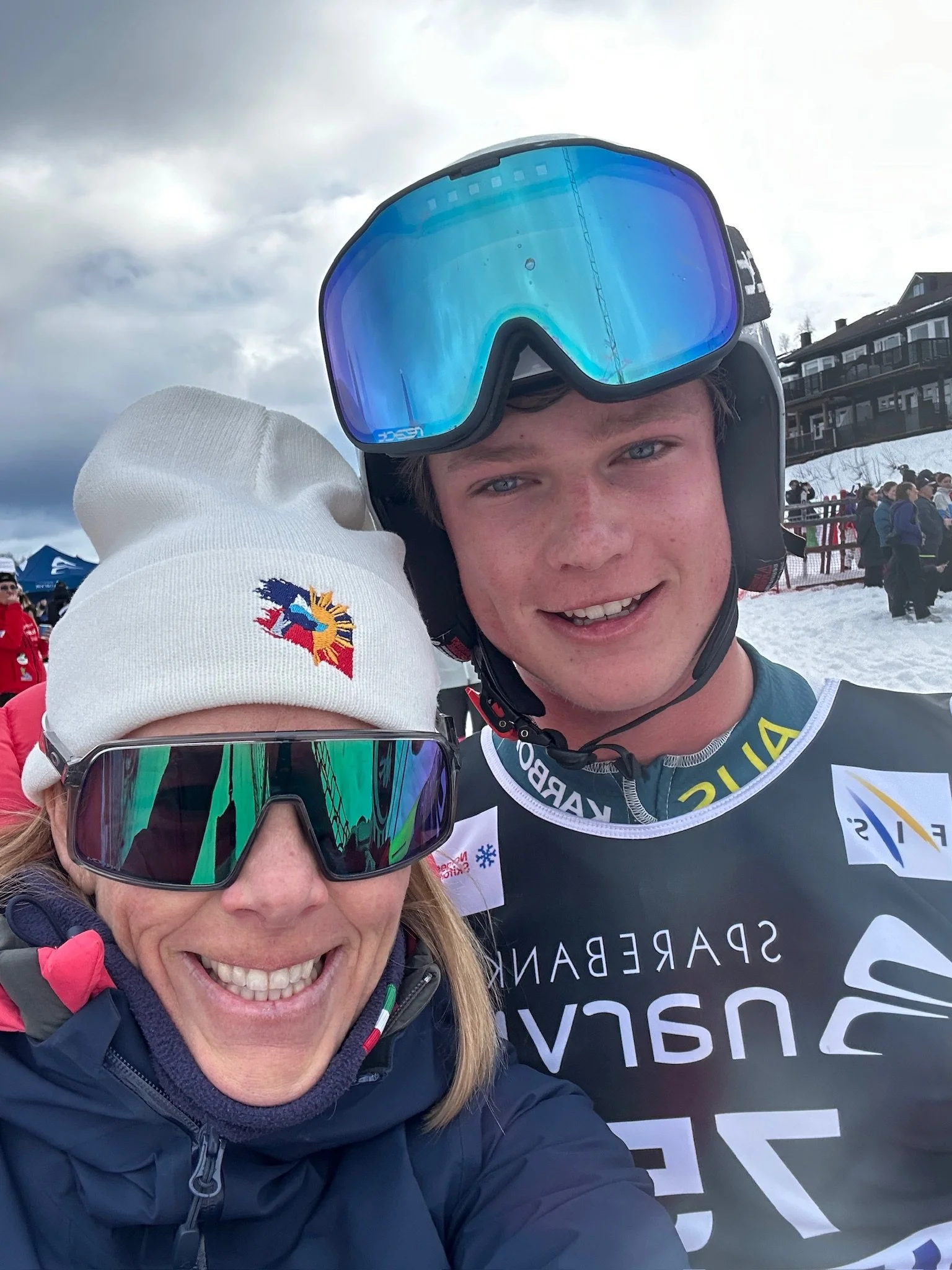 Close-up photo of a smiling young man and woman, both wearing ski gear, standing in snowy terrain during cloudy weather, with a building and a crowd of people in the background.