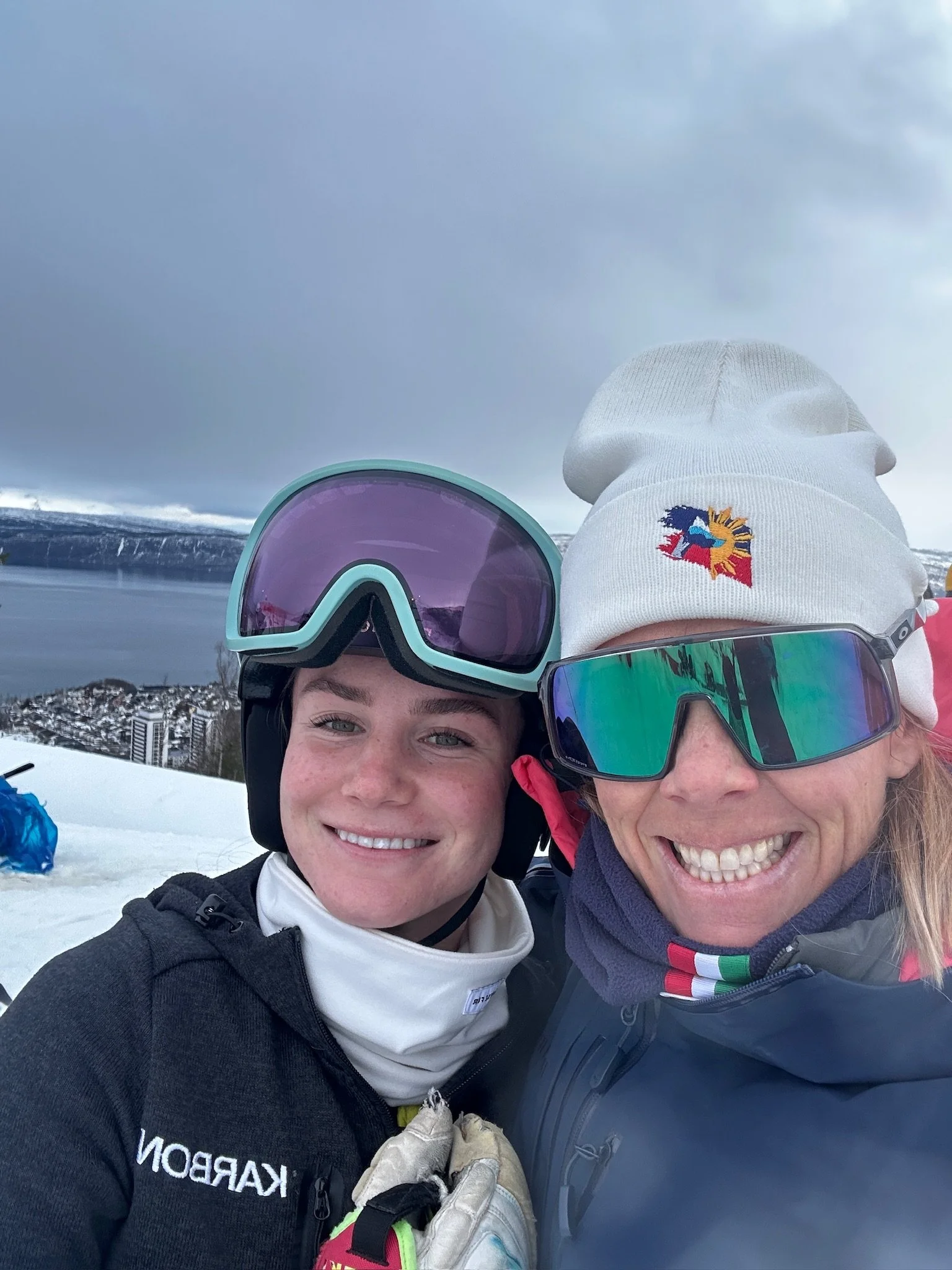 Two smiling people in ski gear taking a selfie on a snowy mountain with a cloudy sky and distant buildings.