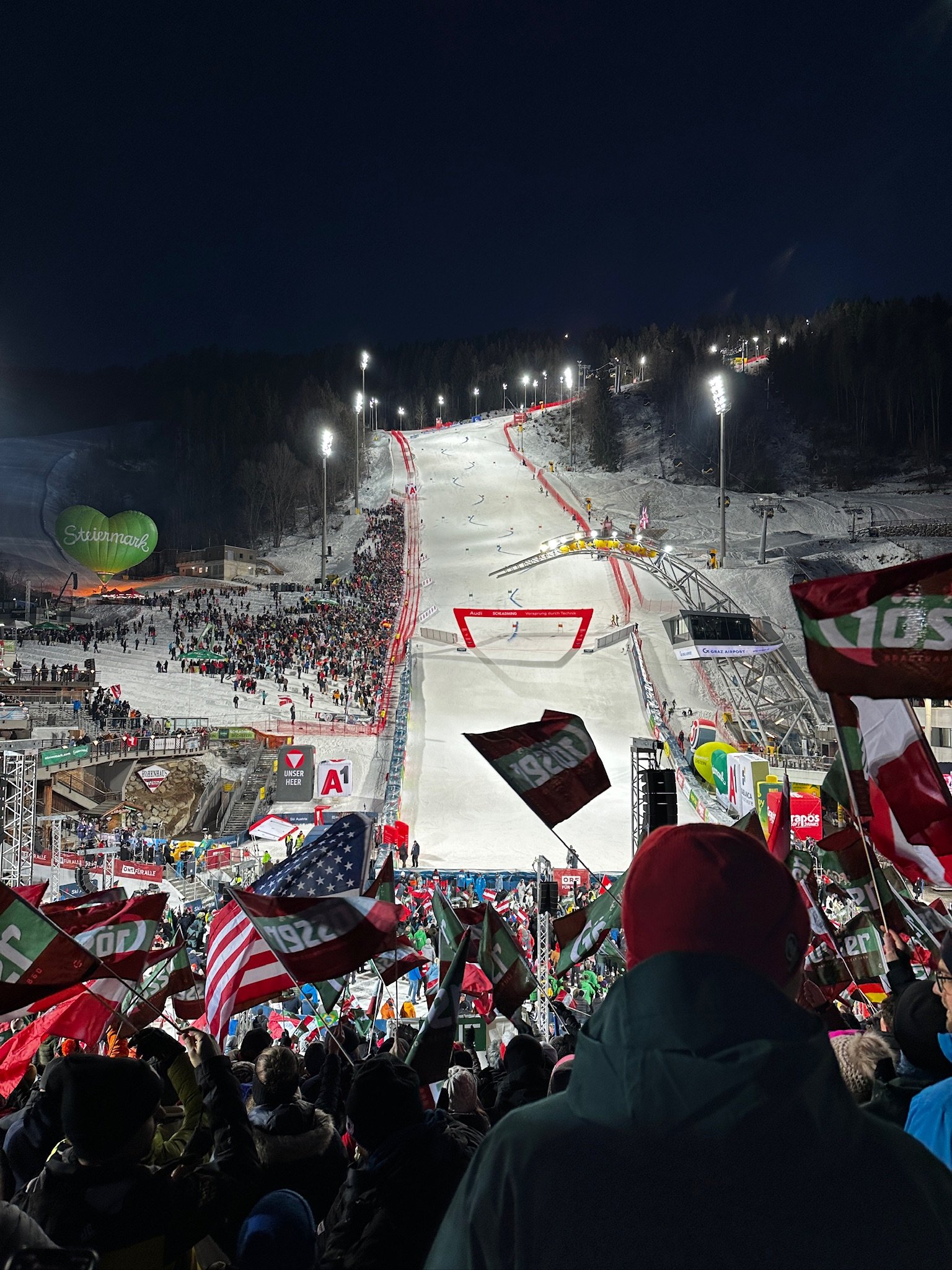 Nighttime at a snowy ski resort with cheering crowd in the foreground, flags, and ski slope with bright lights, chair lifts, and spectators below.