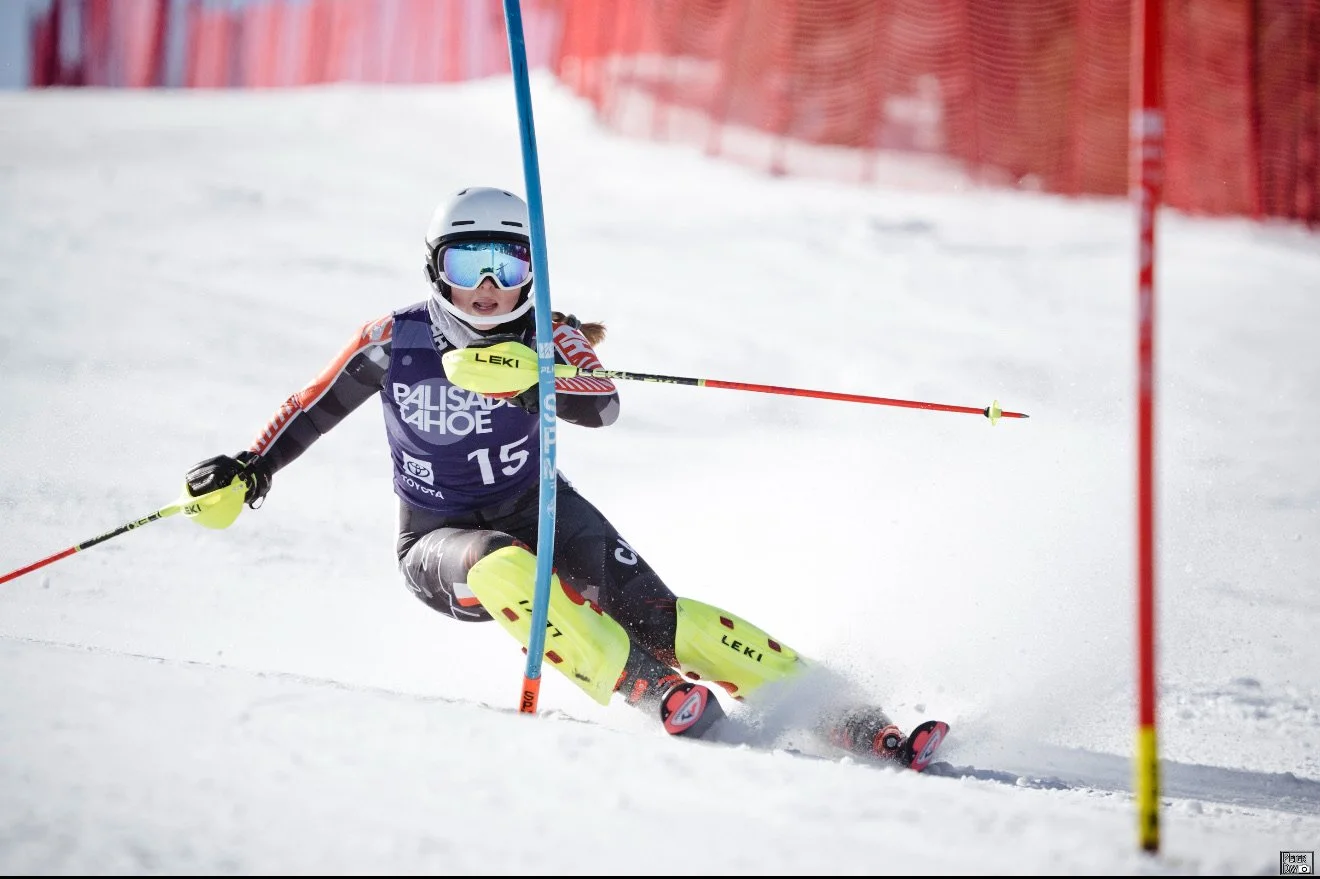 A young skier in protective gear navigates a slalom course on snow, making a sharp turn around a blue and red gate.