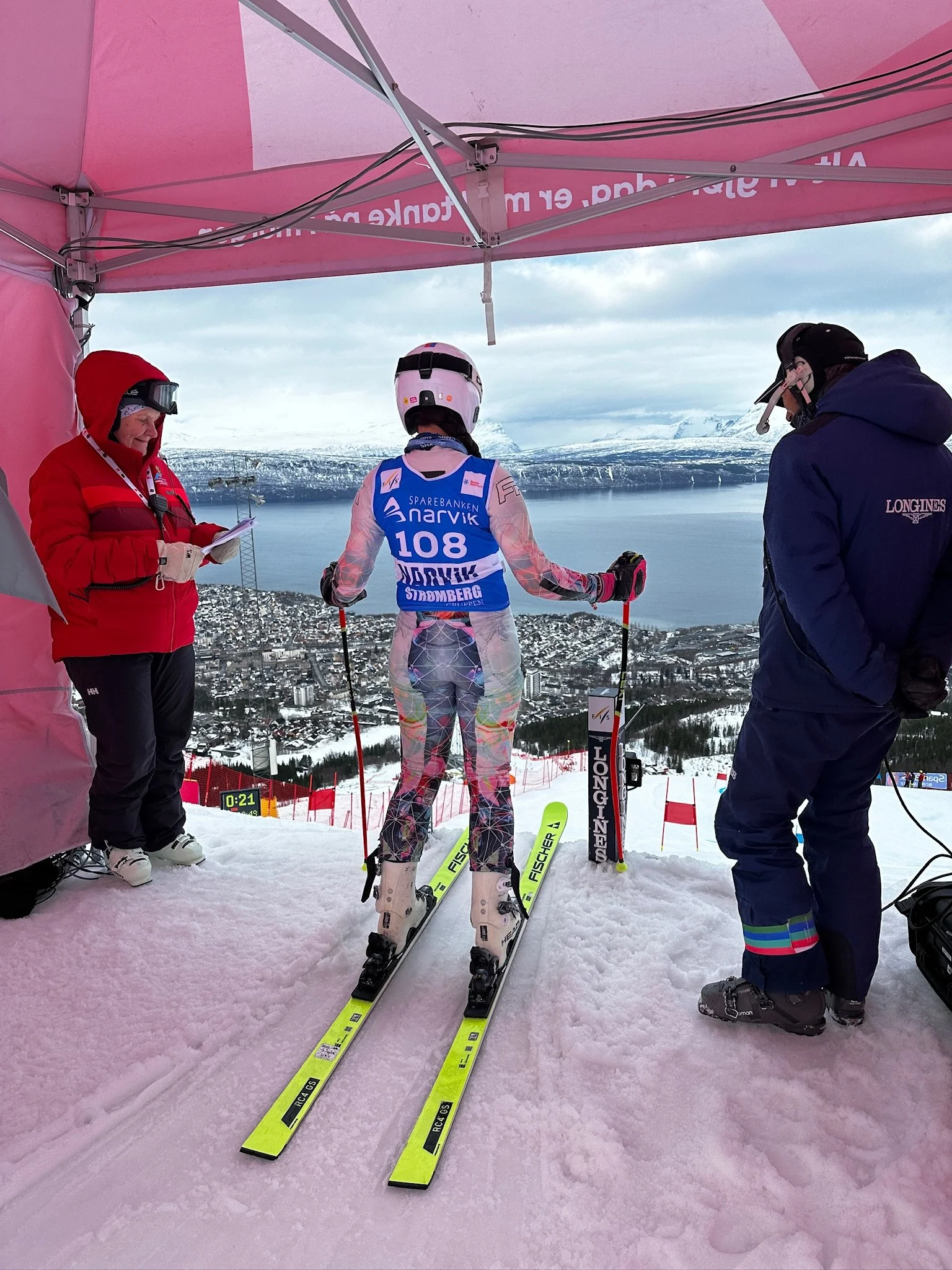 A skier in a floral ski suit with a white helmet and goggles stands on a snow-covered slope, viewed from behind, with two officials or coaches on either side, during a competition or training in a mountainous, snowy landscape with a town and lake in 