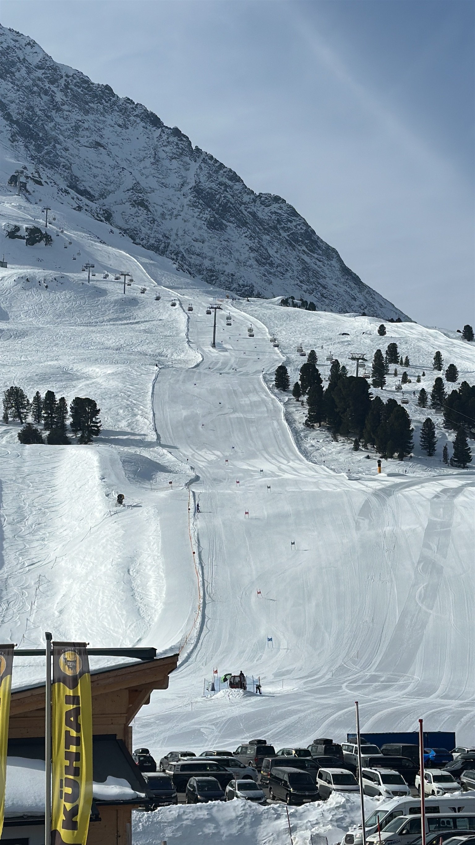 Snow-covered ski slope with ski lifts, trees, and a mountainous backdrop, with a parking lot and building in the foreground.