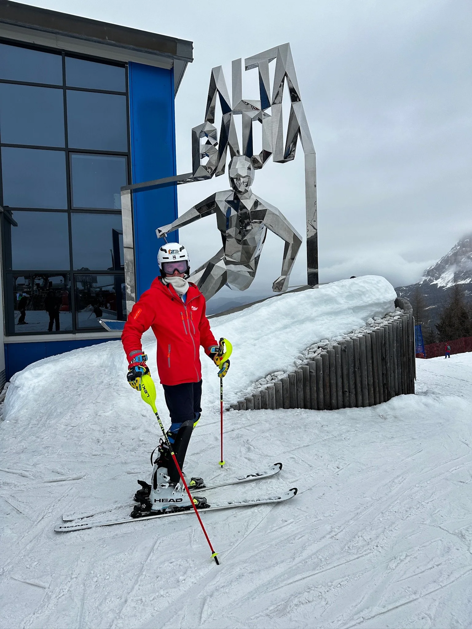 A person wearing a red jacket, black pants, ski boots, and a white helmet with goggles standing on snow with ski poles and skis, in front of a large metallic sculpture of a human figure holding a giant sign with the text 'VIA'. The background shows c